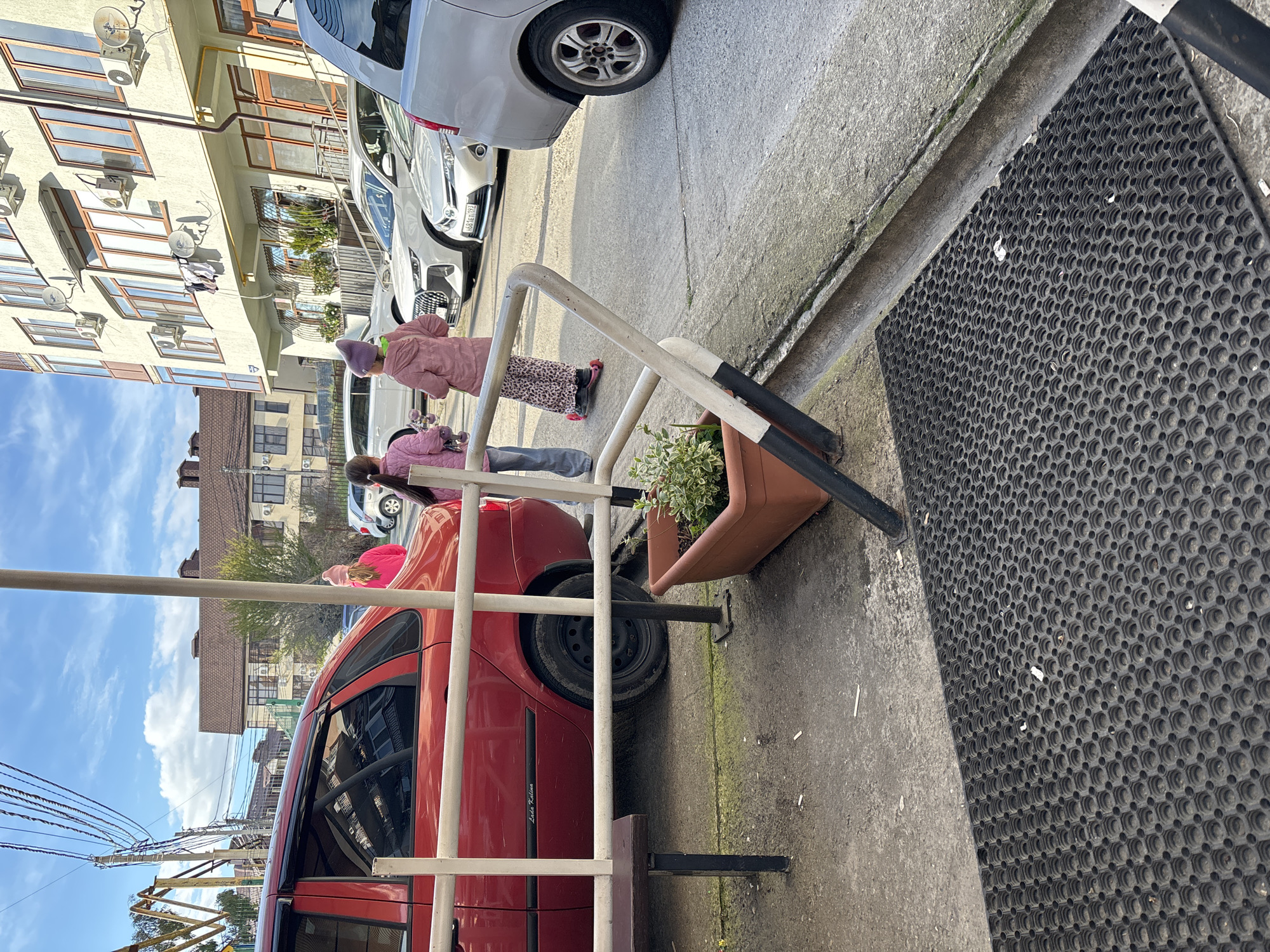 The image shows a street scene in what appears to be Sochi, Russia. Two people are standing near a red car parked along the sidewalk. One person wears a pink jacket and purple hat, while the other has on a pink jacket. A metal railing divides the sidewalk, with a flower pot placed next to it. Behind the red car are other parked cars and buildings with windows and air conditioning units mounted outside. A black rubber mat lies on the sidewalk near the edge of the curb. Overhead, power lines are visible against a blue sky with scattered clouds.