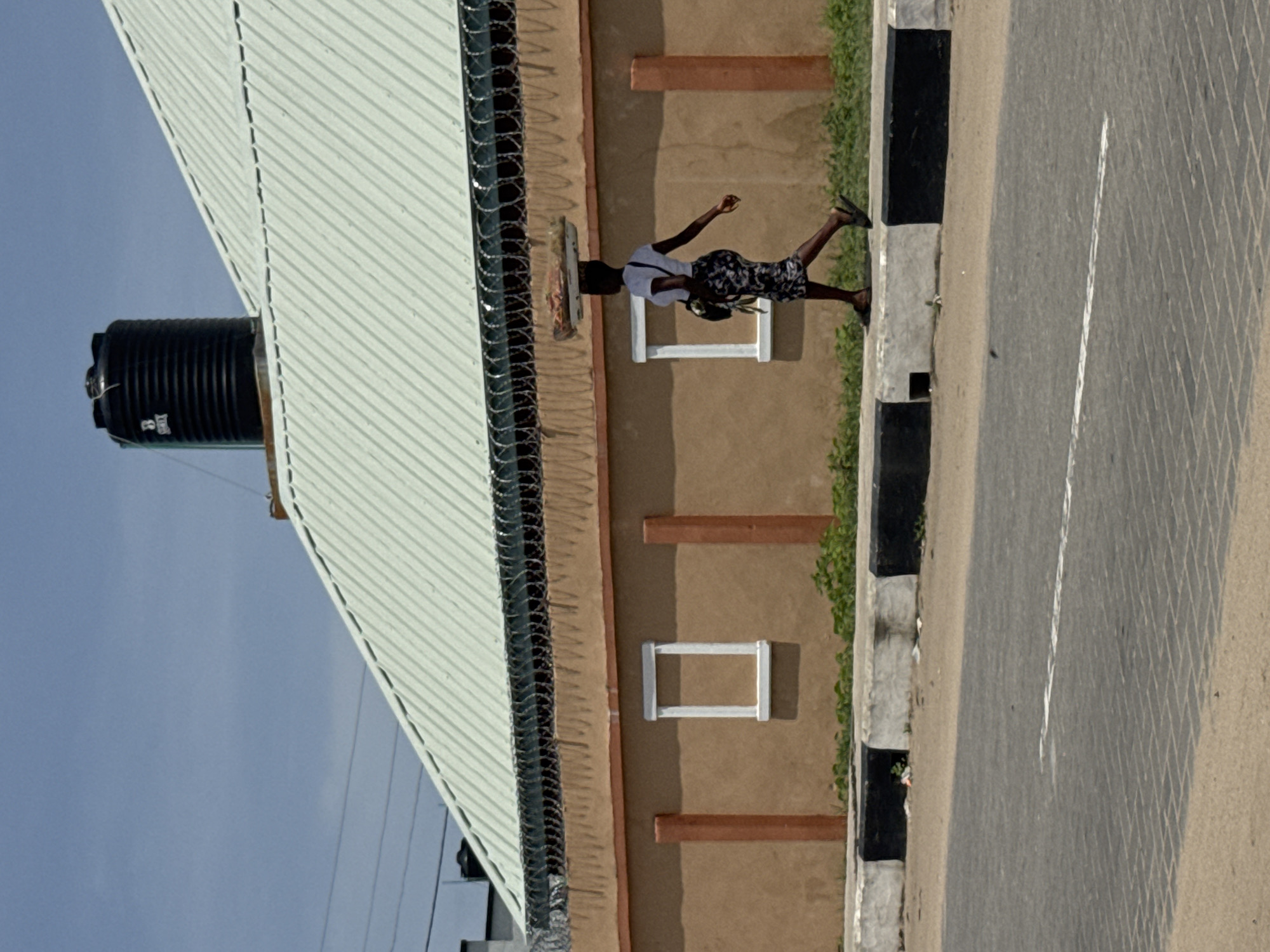 A sideways medium shot depicts a figure walking near a curb along a road in Badagry, Nigeria. The road is asphalt, divided by a white line. Adjacent to the road is a sidewalk with alternating black and white painted posts, alongside greenery. A building sits behind the sidewalk, featuring a light-colored metal roof, a black water tank, barbed wire atop the wall, rectangular architectural details, and a tan-colored wall. The sky is visible.
