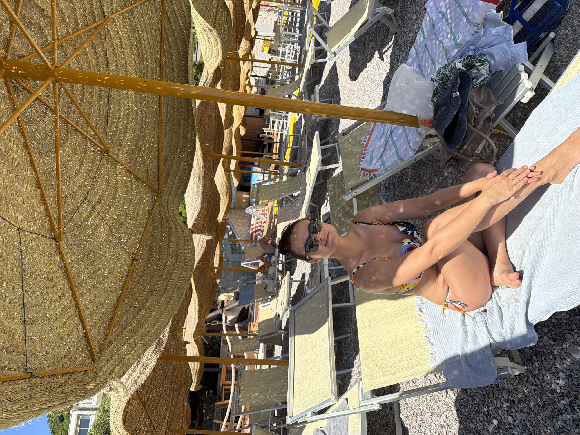 The scene is at a beach in Prijevor, Montenegro. There's a woman wearing sunglasses and a bikini is seated on a beach chair, facing toward the camera. The scene is heavily shadowed by rows of thatch umbrellas. Other beach chairs are visible in the background. Personal items like towels, a hat, and bags are piled on a nearby chair. The ground is gravel.
