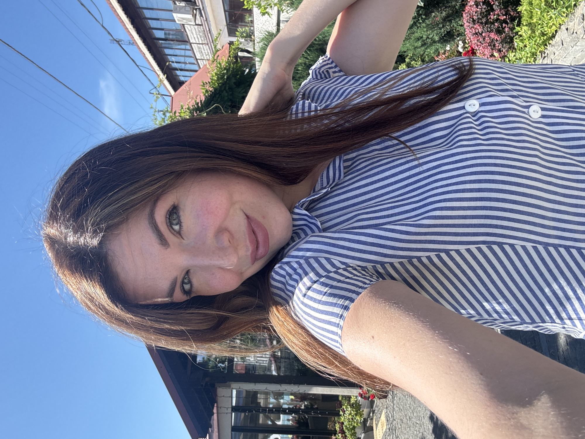 A young woman with long brown hair is taking a selfie outdoors on a sunny day. She is wearing a blue and white striped shirt. Her right arm is bent, and her hand is touching the back of her head. In the background, there is a building with a red roof and some greenery. The sky is clear and blue. The specific location in Odesa, Ukraine, cannot be determined from the image.