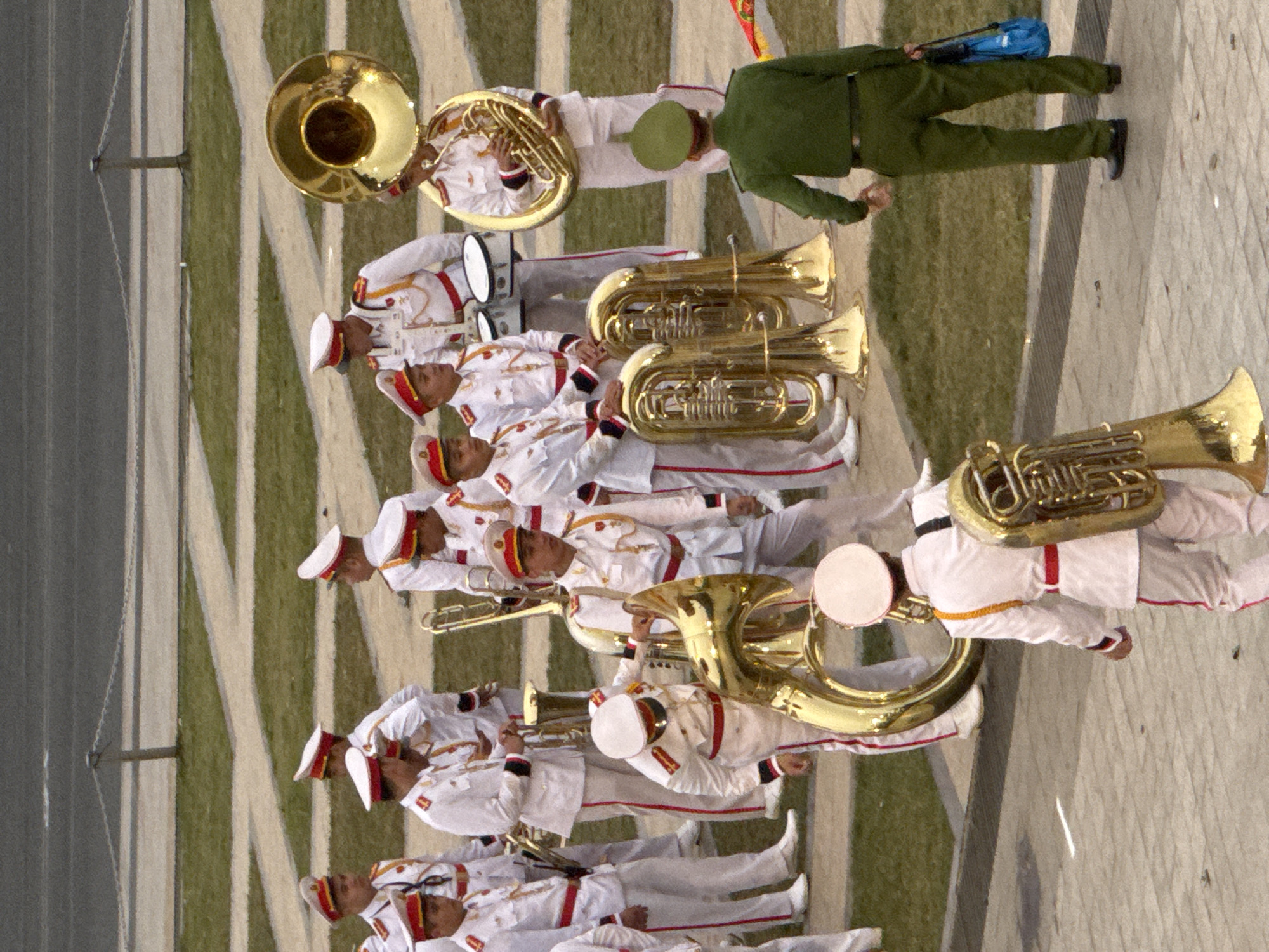 A group of men in matching white ceremonial uniforms with red trim and red-banded hats stand on a paved area with a grassy background. Several of them are holding large brass musical instruments, including tubas and trombones. The instruments are gleaming gold. One man in a green uniform stands facing away from the camera, holding a blue bag. The scene appears to be an outdoor event or performance, possibly a parade or ceremony. The environment suggests a public space, with a paved walkway and a grassy area. The image is shot from a high angle, looking down on the group. Based on the context of similar images, this scene is likely located in the city of Da Lat, Vietnam, possibly at an event such as the Da Lat Flower Festival.