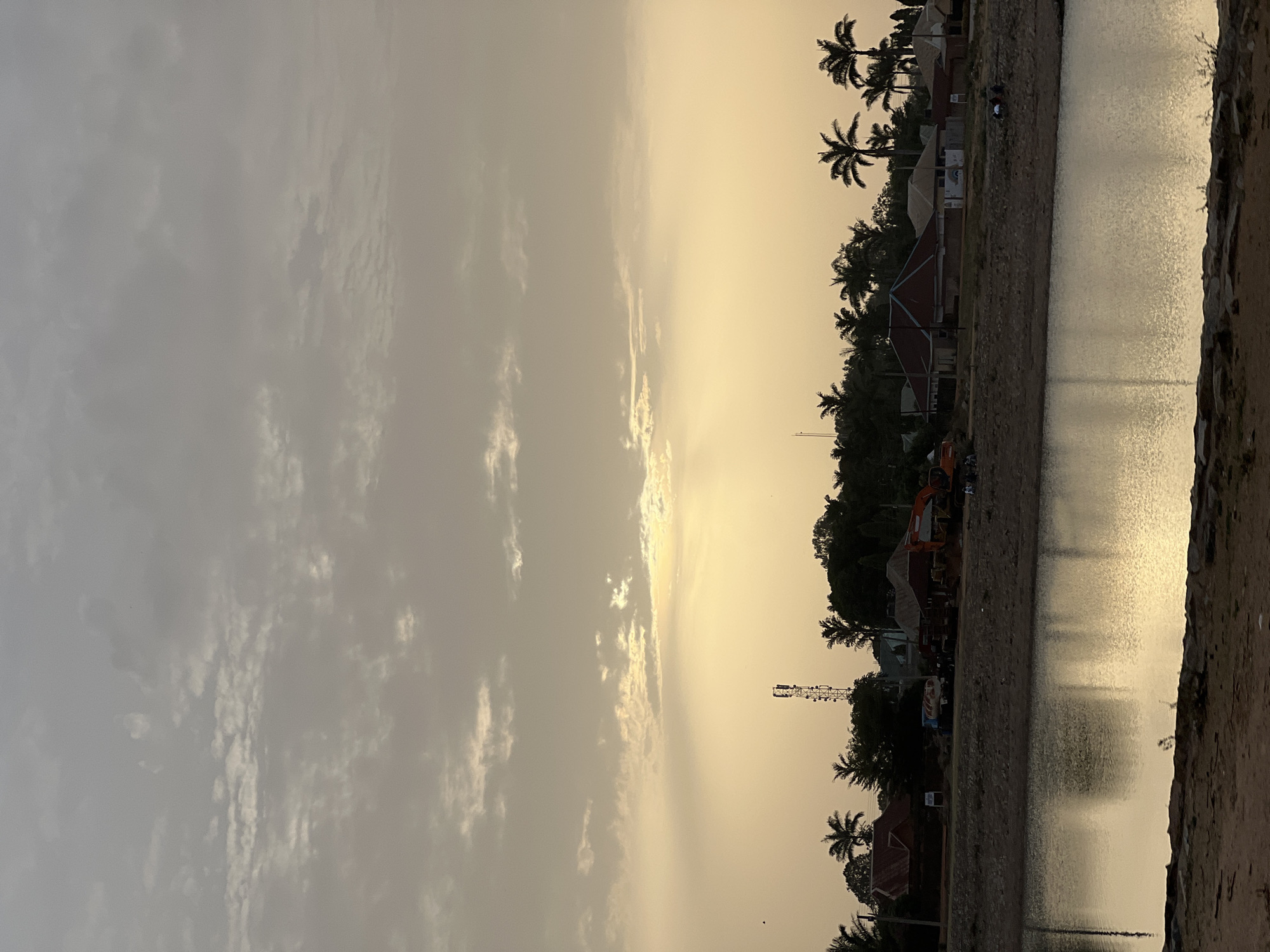 The image depicts a sunset over a body of water in Bukuru, Nigeria. Silhouetted palm trees, buildings, and a communications tower are visible along the shoreline. The sky is filled with clouds, colored with the golden hues of the setting sun. The water reflects the sky, creating a shimmering surface. A construction vehicle is parked near the edge of the water. Some people are near the bank.