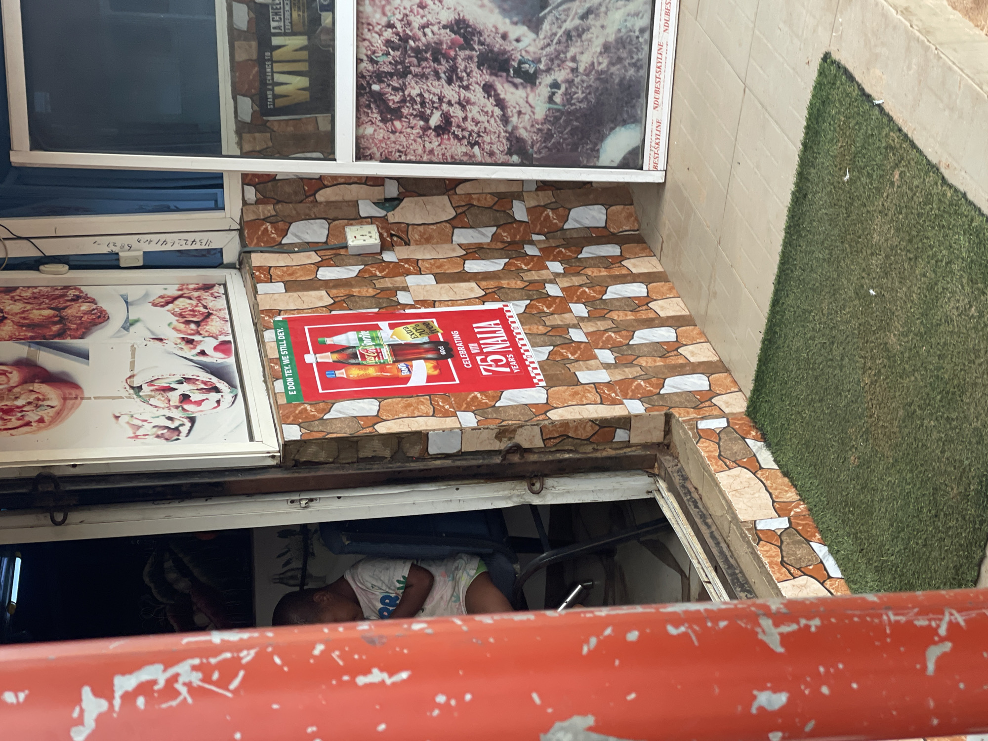The image depicts a street-level view of a small commercial establishment in Lokoja, Nigeria. The facade is decorated with stone-patterned tiles and features a red pillar in the foreground. A poster advertising Coca-Cola and Sprite is prominently displayed on the wall, celebrating "75 Years of Naija." To the right, a glass-paneled door or window reveals an interior with a large image, possibly of food, and text that reads "STAND A CHANCE TO WIN." A young boy is seated inside on a chair, looking down at a device, partially obscured by the open doorway. The ground outside is covered with a patch of artificial grass. The overall scene suggests a typical urban commercial setting in Nigeria, with elements of advertising and daily life.