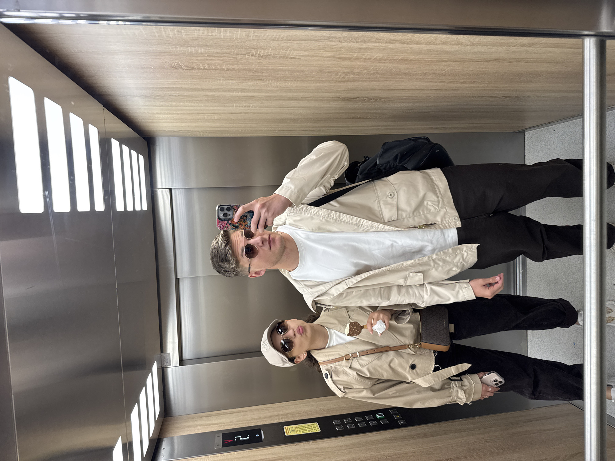 A mirror selfie taken by two individuals in an elevator in Plovdiv, Bulgaria. The elevator features reflective stainless steel walls and light wood-grain panels, with rectangular light fixtures visible on the left wall.
On the left side of the reflection, a male subject wears a beige trench coat over a white t-shirt, dark trousers, and brown sunglasses. He is holding a smartphone with a floral-patterned case to take the picture, and a black backpack is visible behind him.
To his right, a female subject wears a similar beige trench coat over a white t-shirt, dark trousers, a light-colored baseball cap, and brown sunglasses. She holds a chocolate-coated ice cream bar in her right hand and a smartphone in her left. A small patterned crossbody bag hangs from her shoulder. Her lips are slightly puckered in a playful expression.
On the elevator control panel, a digital display shows "2" with an upward arrow. Buttons for floors "1" through "13", "Lobby", and a bell icon are visible. A yellow unreadable informational sticker is also present. The scene suggests daytime, possibly warm weather.