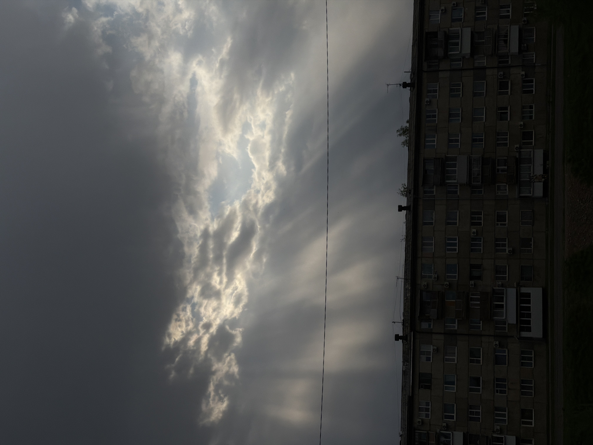 The image depicts a tall building in Birobidzhan, Russia, situated under a sky dominated by dark, ominous clouds with a small break allowing sunlight to penetrate. The building's facade, visible from the side, features numerous windows, some open, suggesting possible ventilation or recent occupancy. Air conditioning units are visible, mounted below some windows. A wire runs across the sky, partially obscuring the clouds. The scene is characterized by low lighting conditions, casting shadows and obscuring details of the building's structure. The presence of sunlight breaking through the clouds creates contrasting beams.