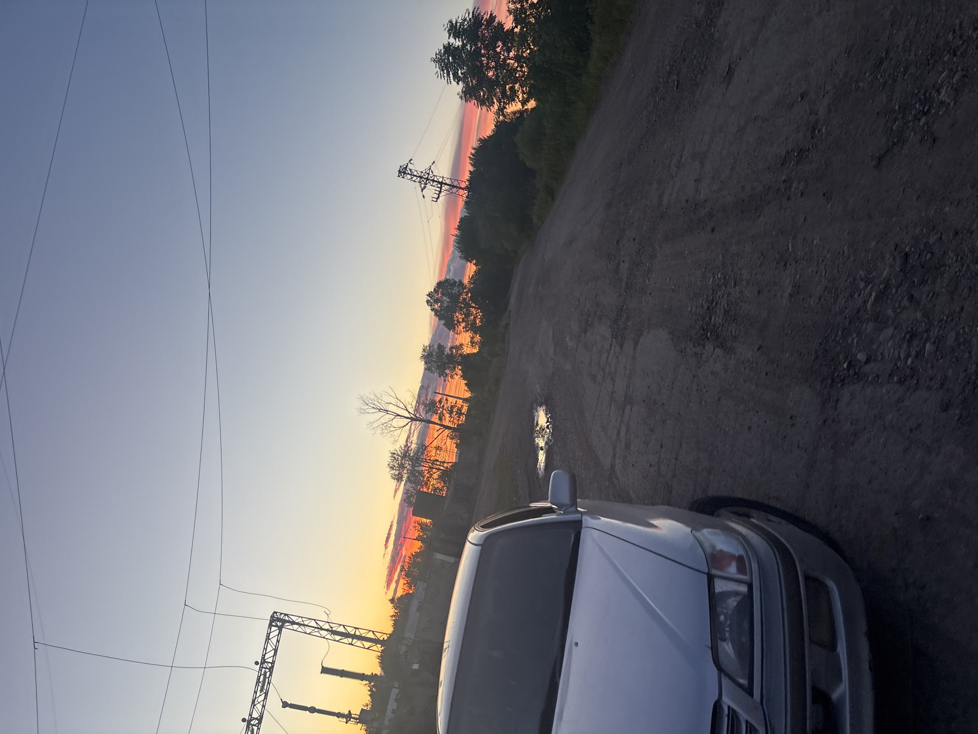 This is a landscape photograph taken at sunset in Birobidzhan, Russia. A silver sedan is parked on a dirt road, partially visible in the foreground. The road is unpaved and shows signs of wear and tear, with puddles of water scattered along its surface. In the background, a power line crosses the frame, supported by metal towers. The sky displays a gradient of colors, transitioning from a soft blue at the top to vibrant orange and pink hues near the horizon, indicative of a sunset. Trees and vegetation line the sides of the road, and a few structures are visible in the distance. The overall scene suggests a rural or semi-industrial setting on the outskirts of the city.