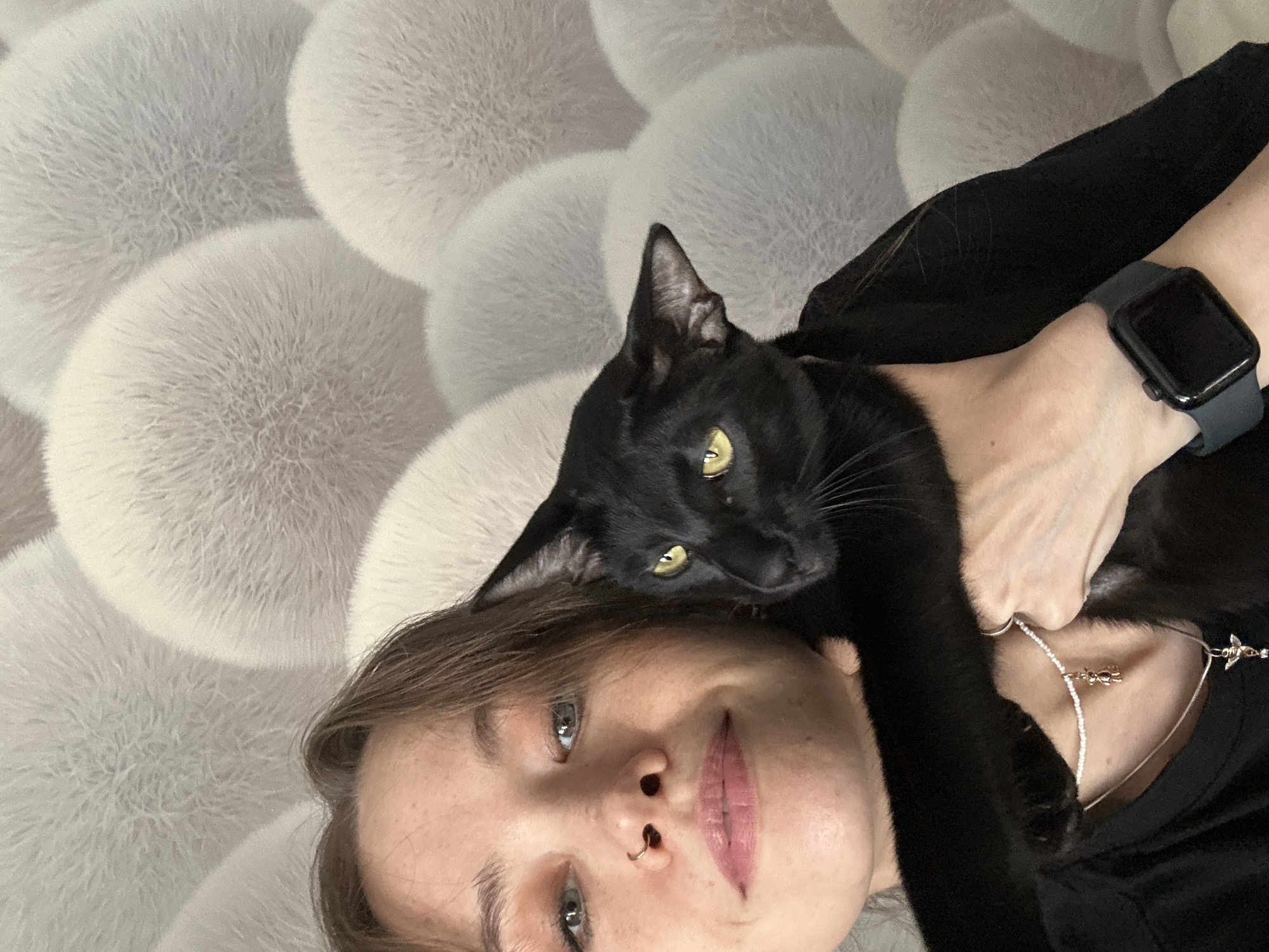 This media file is a selfie featuring a young woman and a black cat. The woman is lying down, with the cat draped around her neck and shoulders. The cat is black with bright yellow eyes, and it is looking directly at the camera. The woman's face is partially visible, and she appears to be looking at the camera as well. She has light brown hair and is wearing light makeup. A silver necklace with a small charm is visible on her neck, as is a dark smartwatch on her wrist. The background is a soft, textured surface that appears to be a fuzzy blanket or rug with a pattern of large, round shapes. The lighting is soft and even, suggesting an indoor setting during the daytime. The overall mood is affectionate and relaxed, with the cat appearing comfortable and content in its position.