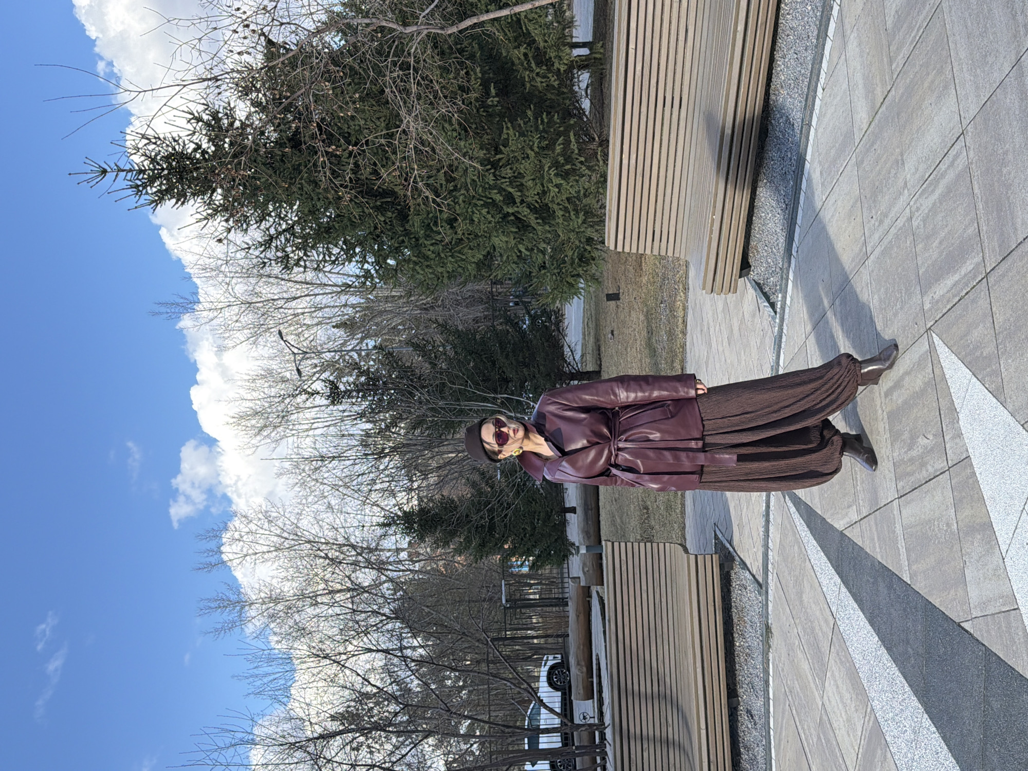 A woman stands posing on a paved outdoor walkway in what appears to be a park or urban plaza in Krasnoyarsk, Russia. She is centrally located in the frame, facing slightly towards the right and looking at the camera. She wears a dark reddish-brown, possibly leather, coat or jacket, cinched at the waist with a belt, paired with wide-leg, pleated dark brown trousers and dark shoes or boots. A dark hat is on her head, and she wears reddish-tinted sunglasses, obscuring her expression.
To her right, two modern wooden benches are visible. The ground beneath her is a light grey paved surface featuring distinct geometric patterns in darker grey and white. In the background, a mix of trees is present; some are evergreen with dark green needles, while others are deciduous with bare branches, suggesting late winter or early spring. A patch of dry grass is also visible beyond the benches. Further in the distance, a white car is partially visible on what seems to be a road or parking area. The sky above is clear and bright blue with scattered fluffy white clouds, indicating sunny weather. The overall impression is a casual outdoor portrait during cool, clear daytime conditions. There is no visible text.