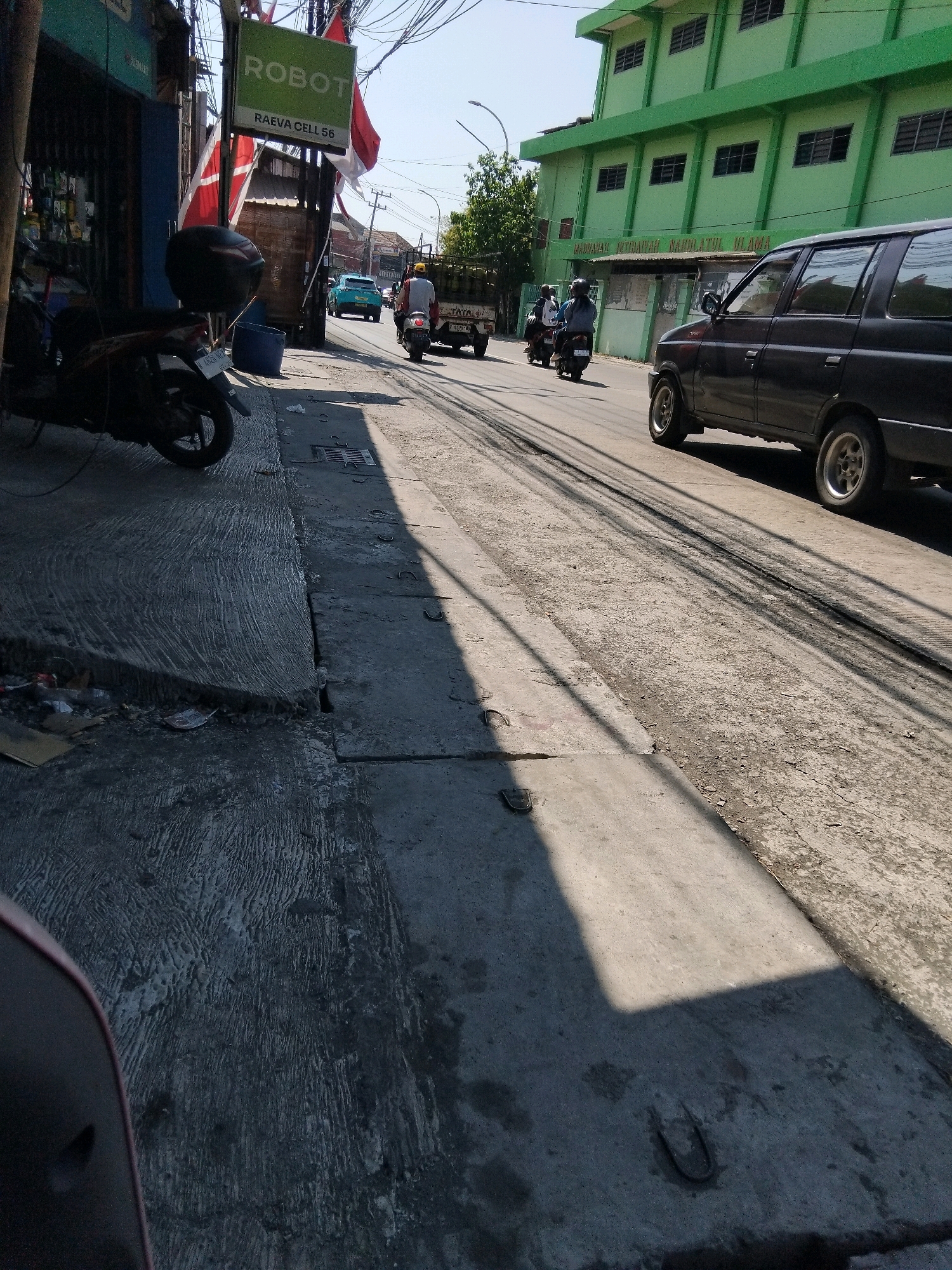 The image shows a street scene in Kepuh Kiriman, Indonesia. A black SUV is parked on the right side of the road, facing away from the camera. To the left of the road, a motorcycle is parked on the sidewalk. A green building with multiple windows is visible in the background. Several people are seen riding motorcycles on the road. The street is paved and appears to be well-trafficked. The lighting suggests it is daytime.