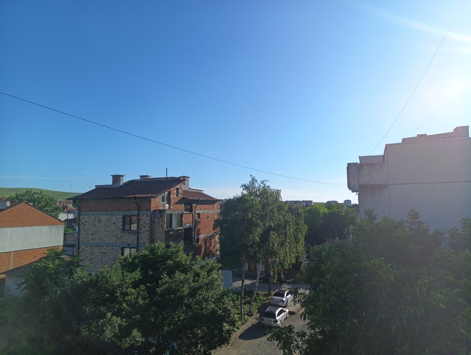 The image depicts an outdoor scene in Karnobat, Bulgaria, under a clear blue sky with bright sunlight and lens flare in the upper right corner. In the foreground, lush green trees obscure a portion of the view. Beyond the trees, two silver sedans are parked on an asphalt road. Further back, there are several buildings. To the left, a brick building with a white facade is visible. Adjacent to it is a multi-story brick building under construction, with exposed brickwork and concrete elements. Another building with a tiled roof and brick facade is situated to its left. In the background, a distant hillside with some vegetation can be seen, along with more buildings and trees, indicating an urban or suburban environment. A thin wire stretches across the sky from left to right.