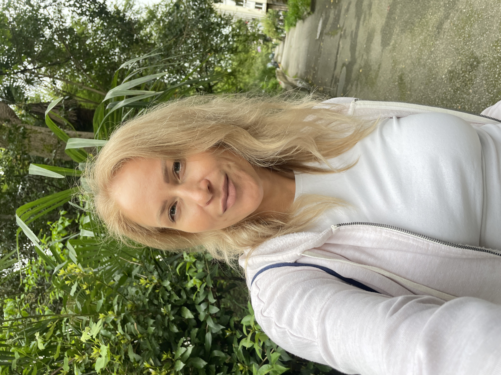 The image is a selfie of a blonde woman in Sochi, Russia. She is wearing a white t-shirt and a light pink hoodie with a blue strap, with a blurred background of a path, greenery, and a building, all indicating an outdoor setting.