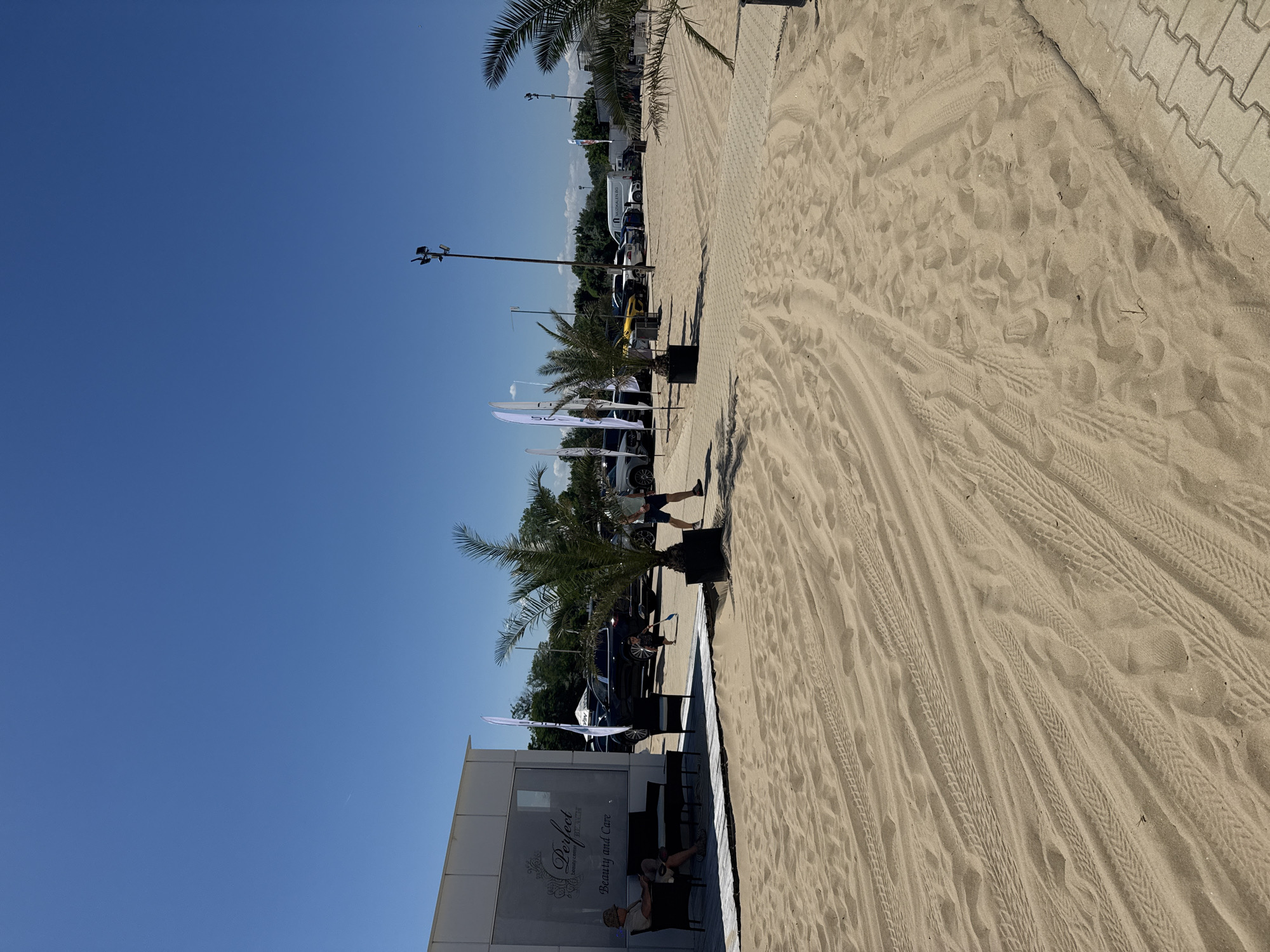 This image depicts a beach scene in Nesebar, Bulgaria, on a clear, sunny day. The foreground is dominated by sand with visible tire tracks, suggesting vehicle activity on the beach. To the left, a white building with a sign reading "Perfect Beauty Center Beach" is partially visible, with two people seated on chairs outside. In the background, several cars are parked, and palm trees are scattered throughout the area. A few white flags are also visible, potentially indicating event signage or branding. The sky is a vibrant blue, with no clouds present.
