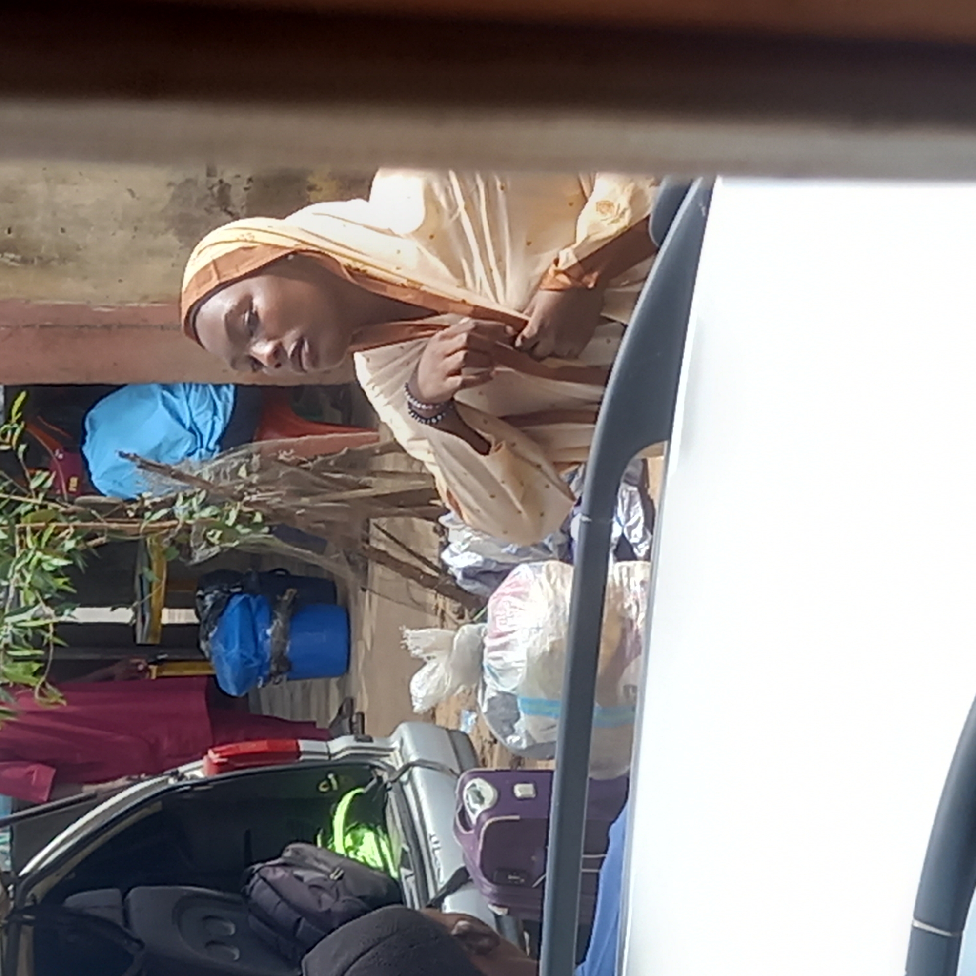 The image depicts a woman with dark skin, wearing a light-colored headscarf and clothing with brown trim. She is looking down and slightly to the left, her hands resting on her lap. She is standing near the open trunk of a car filled with luggage and other belongings, suggesting a possible relocation or travel scenario. The setting appears to be outdoors, with some vegetation and a wall in the background. The location is potentially in the city of Dadi Riba, Nigeria, based on provided information.