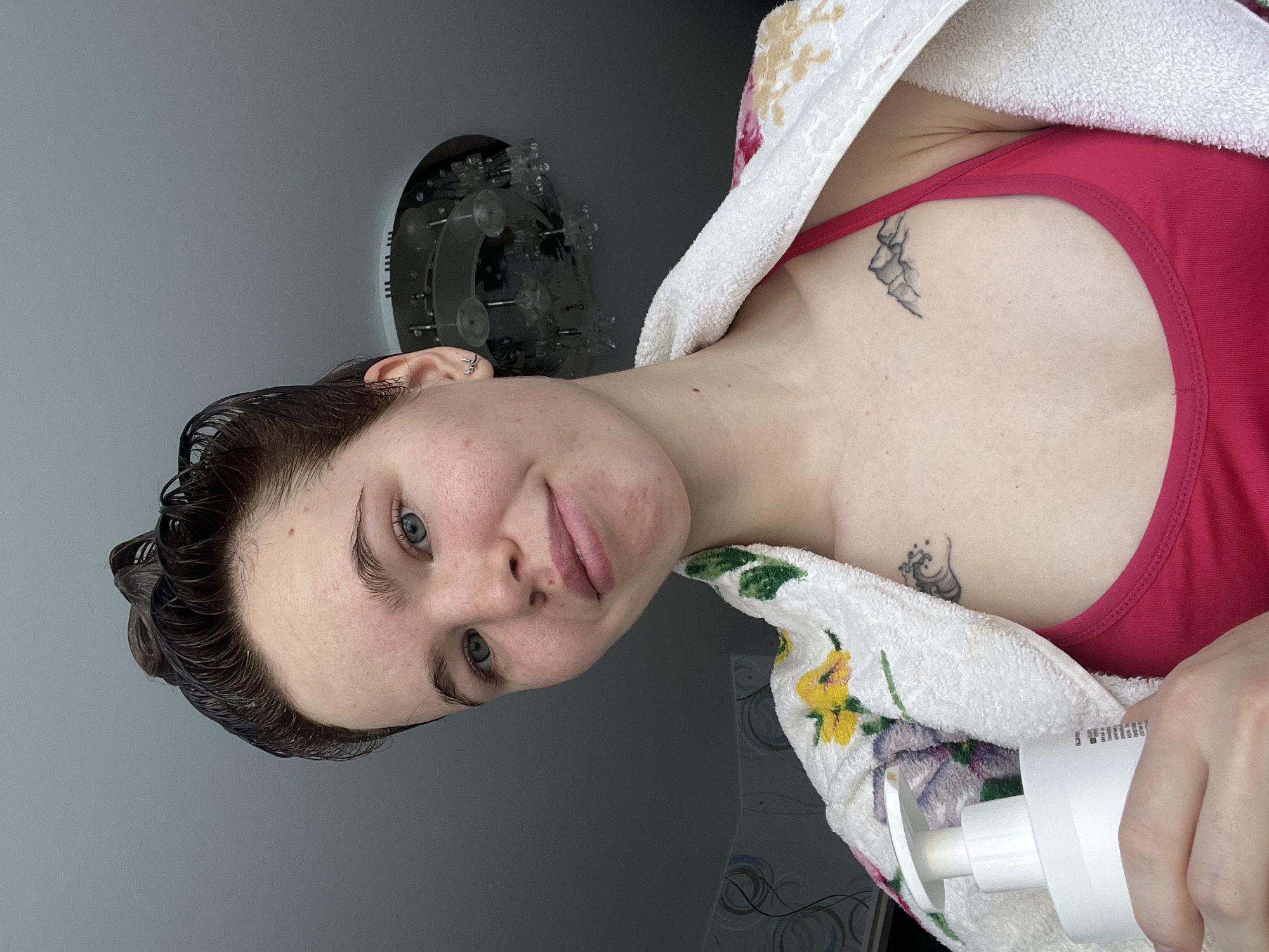 A woman with wet dark brown hair, light skin, and blue eyes is facing the camera with a slight smile. She is wearing a red tank top and has a white towel with floral patterns draped over her shoulders. Two small tattoos are visible on her chest/collarbone area. She is holding a white bottle with a pump in her right hand.
In the background, a gray wall is visible, along with a decorative lighting fixture on the wall. Based on metadata (not visible), the image was taken in Chaikovsky, Russia.
