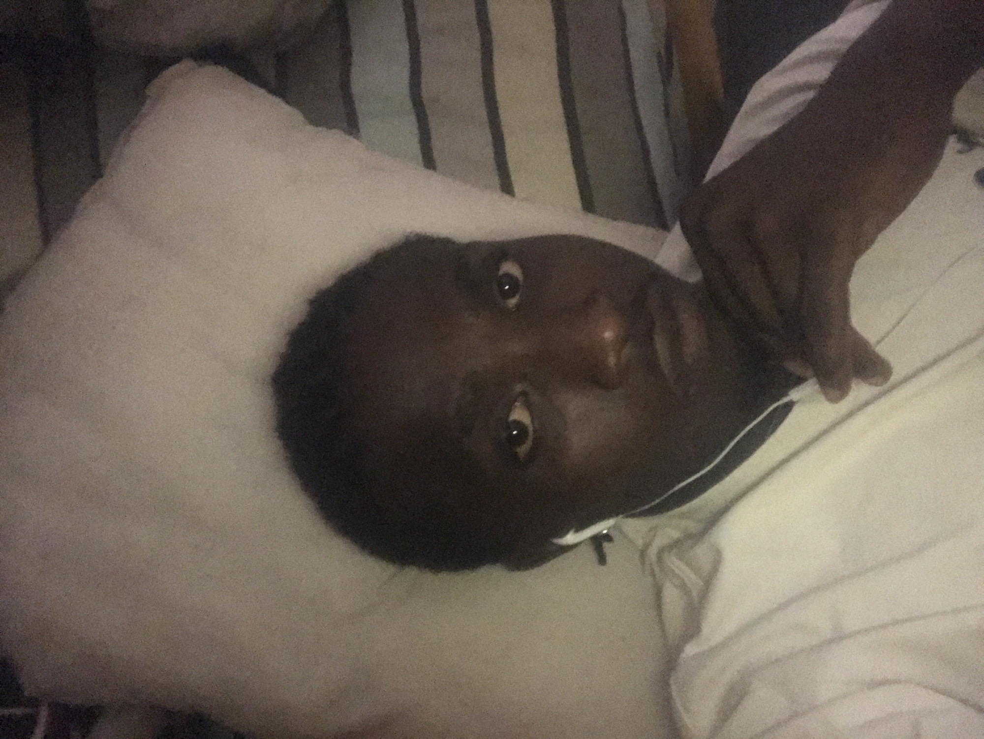 A dark-skinned individual, appearing male, is lying down indoors, looking directly at the camera. The person has short, dark hair and is wearing a white t-shirt and white wired earbuds in both ears. Their right hand is positioned near their chin. A white pillow supports their head, and striped bedding (blue/grey and white) is visible to the right of the pillow. The lighting is dim, contributing to a slight graininess in the image. The setting appears to be a bedroom, specifically a bed. The location is in the city of Unknown, Unknown.