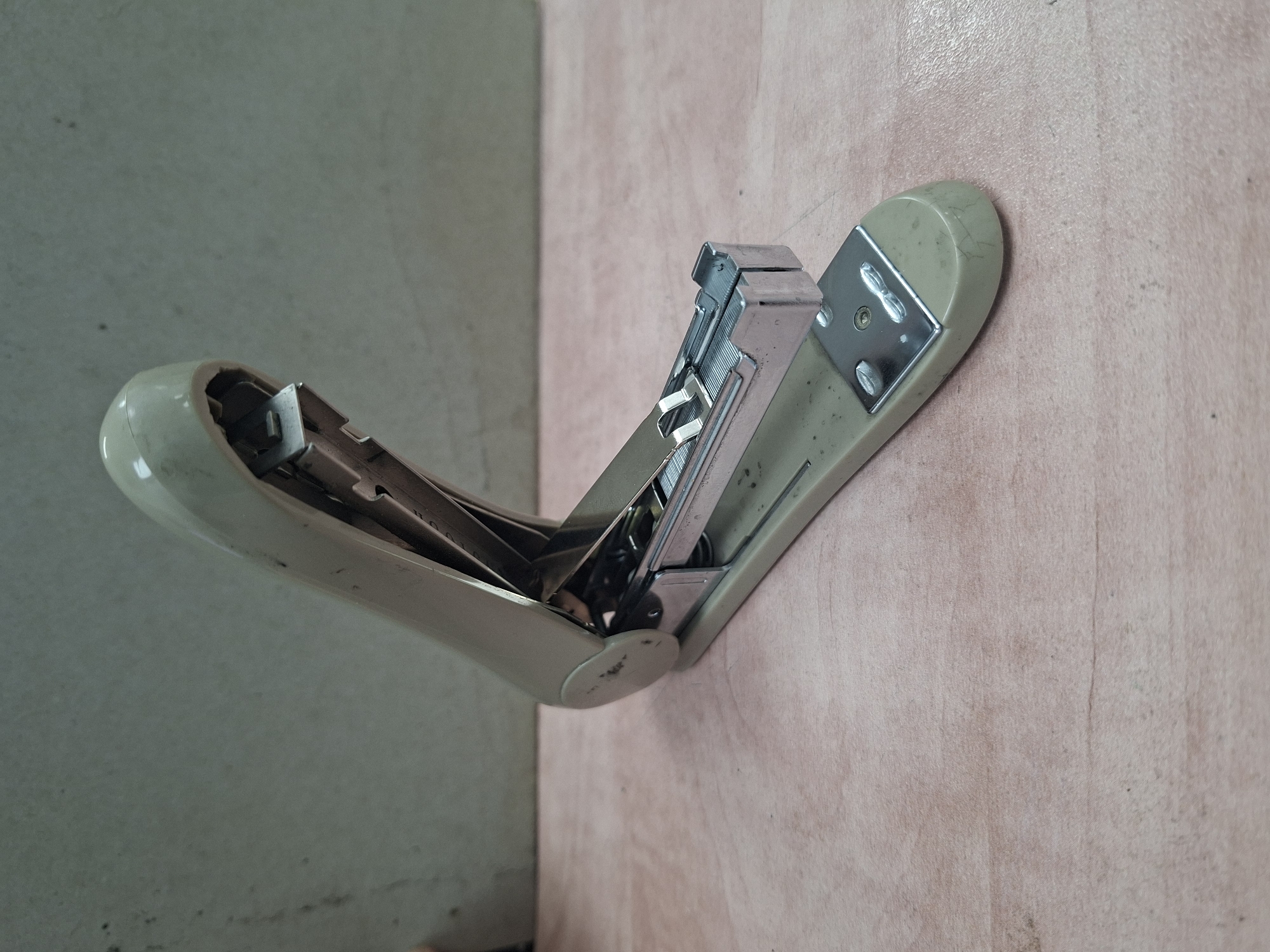 A cream-colored stapler rests on a light pink wooden surface. The stapler is open, revealing its metallic inner workings and a row of staples. The background is a textured, grey wall. The image is taken from a slightly elevated angle, focusing on the stapler. There are no people or animals present in the scene. The environment appears to be indoors, possibly an office or workspace.