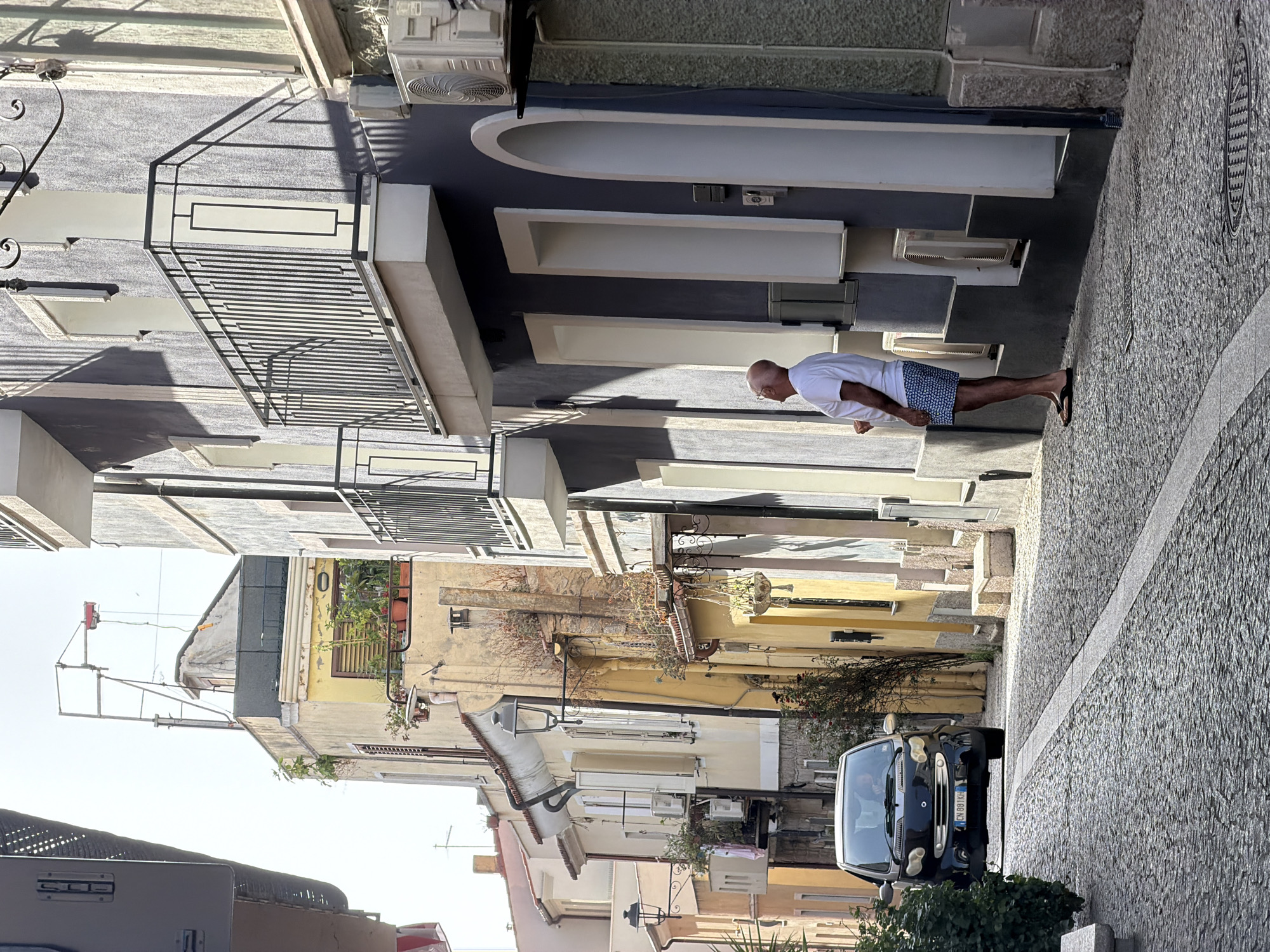In Olbia, Italy, on a sunlit day, a man stands in a cobblestone alleyway, looking upwards at a building. He is wearing a white t-shirt and patterned shorts, with flip-flops on his feet. To his left, a black Smart car is parked. The alley is lined with buildings of various colors, including yellow and grey. Some buildings have balconies with metal railings and air conditioning units. There are also street lamps and vegetation visible. The sky is clear and bright.