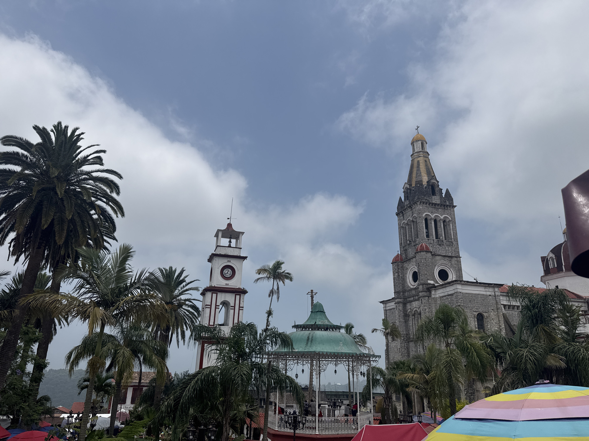 The image depicts a square in Cuetzalan, Mexico, under a partly cloudy sky. In the foreground, there are several palm trees and a colorful striped umbrella. In the middle ground, there's an ornate gazebo with a green roof. Behind the gazebo, two prominent buildings dominate the scene: a white clock tower with red trim and a stone church with a tall steeple. The clock tower has clock faces on multiple sides, and the church steeple is topped with a golden cross. People are visible inside the gazebo. In the background, a blurred mountain range can be seen.