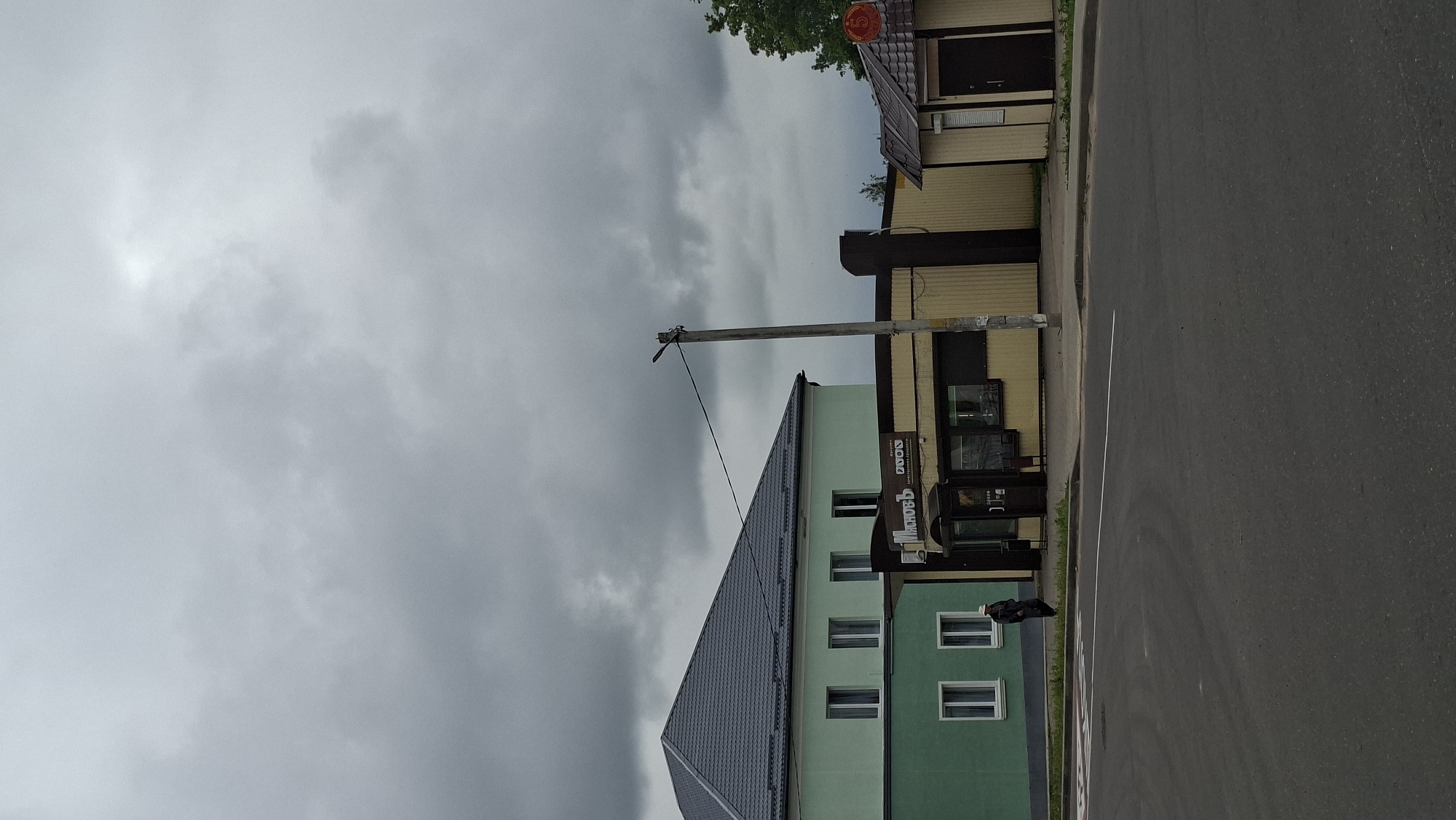 The image shows a street scene in the city of Smalyavichy, Belarus. On the left is a two-story building painted green with a dark grey roof. It has multiple windows on both floors. To the right of the green building is a smaller, light yellow building with a brown trim and a dark brown door. Next to that is a storefront with a yellow facade, dark brown pillars, and large glass windows. A person wearing dark clothing and a light-colored hat is walking on the sidewalk in front of the green building. A utility pole with wires is visible in front of the storefront. The sky is overcast with grey clouds. The foreground shows a paved road with a pedestrian crossing marking.