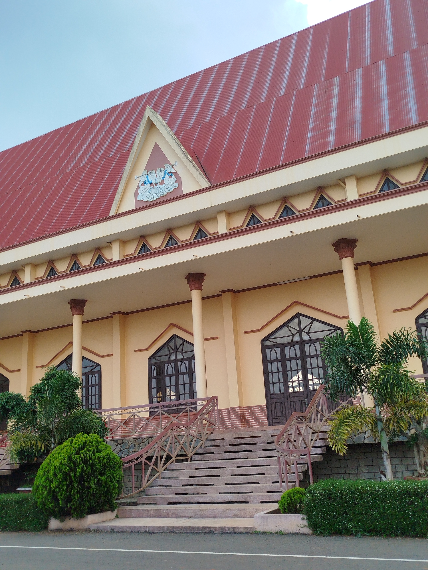 The image displays the exterior of a large, grand building under a clear, light blue sky. The most prominent feature is its expansive, steeply angled red corrugated metal roof, which dominates the upper portion of the frame. At the peak of the roof, a triangular pediment showcases a light blue and white relief sculpture depicting two winged figures, possibly angels or cherubs, blowing trumpets amidst cloud-like elements.
The building's facade is painted in a soft, light yellow, contrasting with dark brown, arched windows and double doors that feature intricate grid patterns. A series of robust, light yellow columns with decorative dark brown capitals support a wide overhang that extends across the front of the structure. Below the roofline, a frieze-like architectural detail features a repeating pattern of small triangular cutouts.
Wide concrete steps lead up to the main entrance, flanked by sturdy wooden railings with a distinctive geometric design, supported by bases of brick and stone. The immediate surroundings are landscaped with lush green vegetation, including neatly trimmed bushes and several tall palm trees that add a tropical touch to the scene. In the foreground, a paved road or driveway with a white line marking its edge is visible. The scene is devoid of human activity, presenting a static view of the imposing and well-maintained building, likely a public or religious structure, during daylight hours in Krái 2, Vietnam. No visible text is present.