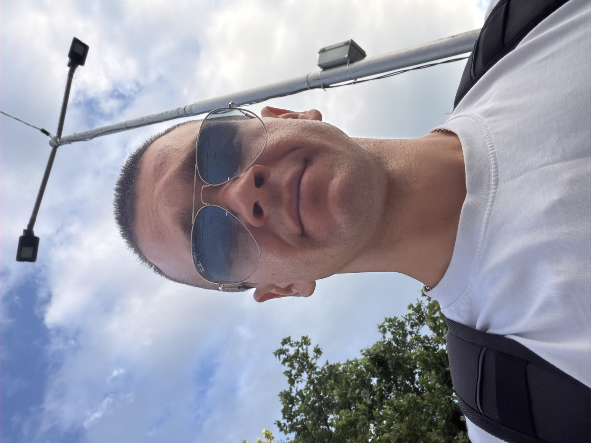 The image is a selfie taken from a low angle outdoors. A person wearing a white t-shirt and aviator sunglasses is looking directly at the camera. They have short hair and are wearing a silver chain. A black backpack is visible on their shoulders. In the background, there is a utility pole with a lamp and a small gray electrical box attached to it, as well as a lush green tree on the left. The sky is cloudy. The setting appears to be a public outdoor space. Based on the provided information, this location is in Veliko Tarnovo, Bulgaria.