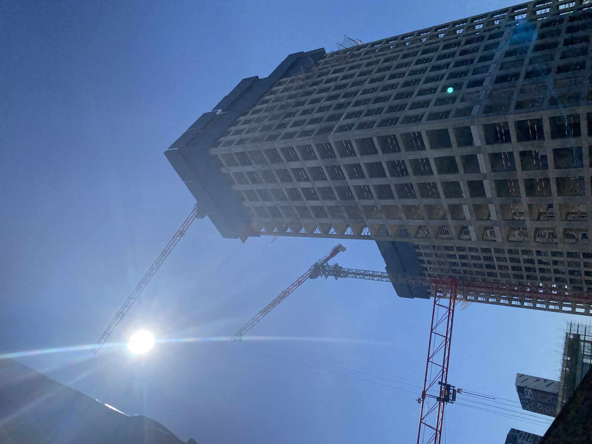 This image shows the construction of a high-rise building in Moscow, Russia, under a clear blue sky. Two construction cranes are visible, one taller and more prominent than the other, reaching towards the sky. The unfinished building is made of concrete, with visible floors and window openings. The sun is shining brightly, creating lens flare and casting rays across the scene. The overall impression is one of ongoing urban development.
