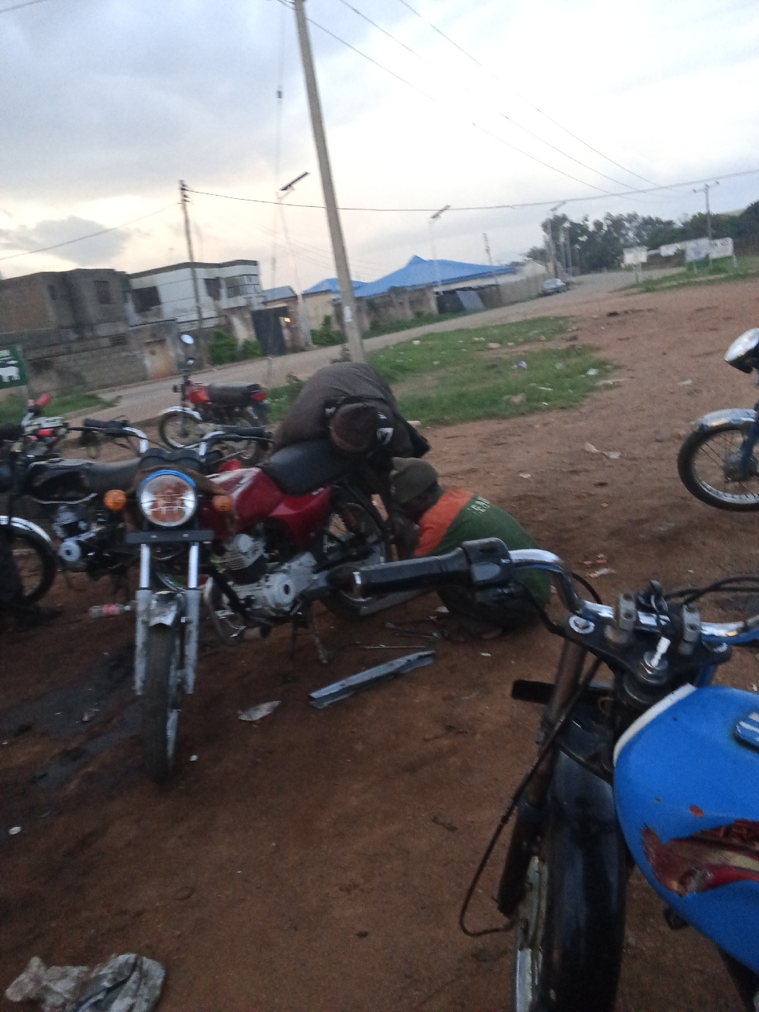 In Barakin Lerawi, Nigeria, the scene depicts an outdoor repair area with multiple motorcycles. Two individuals are actively engaged with the motorcycles. One person, wearing a dark brown jacket and a brown cap, is bent over a red motorcycle, appearing to be working on it. Another individual, dressed in a green and orange vest, is also crouched near a motorcycle, with their hands obscured, likely participating in the repair. Several motorcycles are parked close together, including a red one in the foreground, a blue one with damage to its fuel tank, and other motorcycles further in the background. The ground is a mix of dirt and patchy grass. In the background, buildings with corrugated iron roofs and concrete structures are visible, along with overhead power lines and a utility pole. The sky is overcast, suggesting it may be late afternoon or early morning. The overall impression is of a functional, informal motorcycle repair or maintenance area.