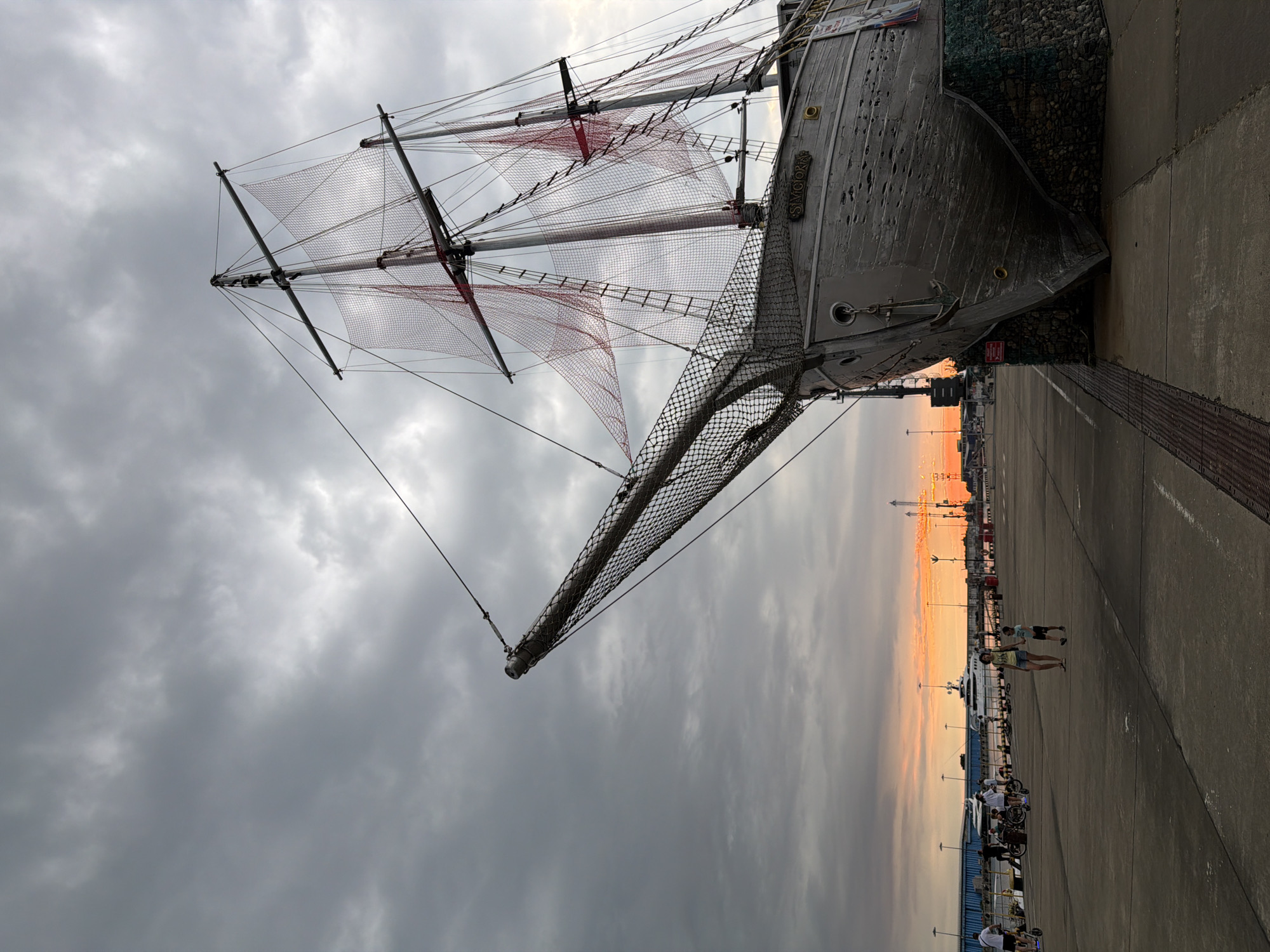 This image captures a section of the Sirius city waterfront in Russia, dominated by the bow of a large, stationary wooden sailing ship sculpture. The ship, named "SILVICTORIA" as indicated by lettering on its hull, is propped up on a stone base. Its masts are visible, adorned with red netting. In the background, the sky is overcast with dramatic clouds, but a vibrant sunset is breaking through on the left, casting an orange glow on the horizon. The waterfront promenade in front of the ship is paved with concrete. Several people are present on the promenade: a woman and a young boy walk together in the mid-ground, while further back, individuals are seen on electric scooters and bicycles near the water's edge, where several yachts are moored. The overall atmosphere suggests a late afternoon or early evening scene at a popular public space.