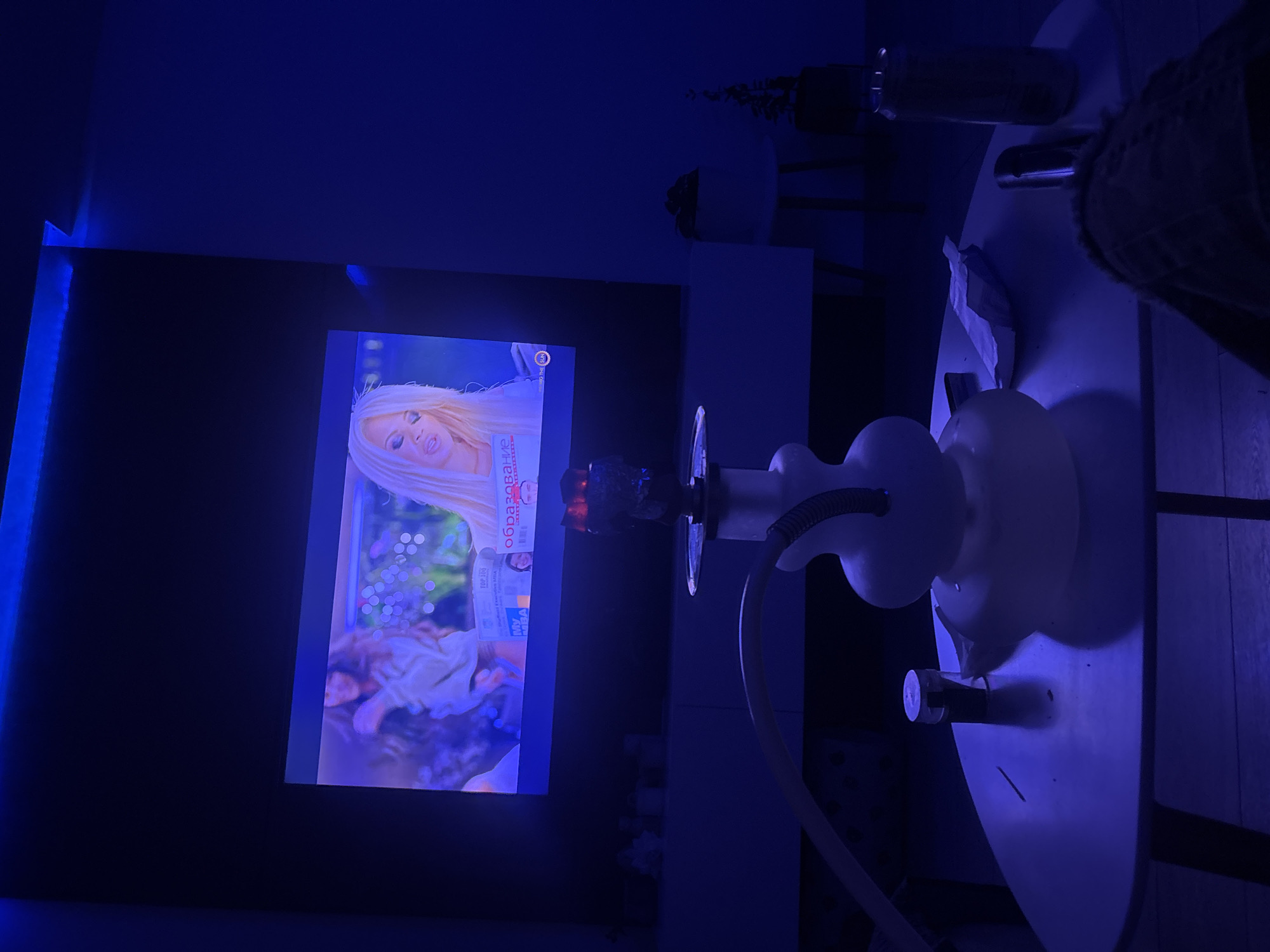 In an indoor environment with blue ambient lighting, a hookah is positioned on a coffee table. The hookah features a white base and a clear hose. On the table, there are also a small cylinder, a crumpled paper item, and a can. In the background, a television screen displays an image of a woman with blonde hair, and a thin blue light strip is mounted above the television. The scene appears to be a living room.