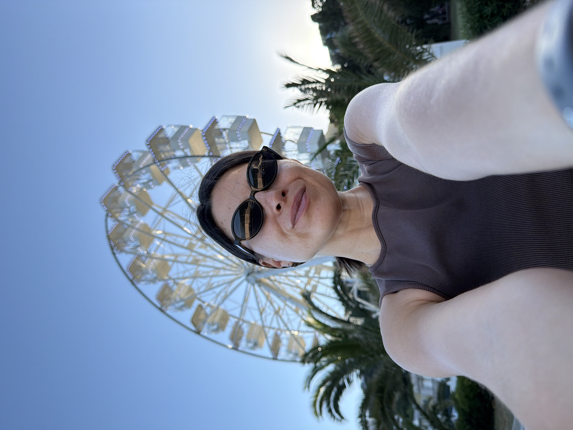 A female subject, appearing to be in her late 20s to early 40s, takes a selfie, holding the camera with extended arms. She wears dark, oval-shaped sunglasses and a dark brown ribbed sleeveless top. Her dark hair is pulled back. A faint smile is visible on her lips. A dark band or bracelet is visible on her right wrist (viewer's left).
In the background, a large, white-framed Ferris wheel stands prominently, slightly out of focus. Its cabins are visible, and some elements of its structure are illuminated with small, possibly blue or white, lights. Palm trees flank the scene on both the left and right sides of the subject, with their fronds partially obscuring parts of the Ferris wheel and the distant background. The sky above is clear and bright blue, suggesting daytime or early evening, with a subtle warmer hue on the right side indicating a low sun angle. Distant buildings are vaguely discernible behind the palm trees. The scene depicts a leisure or resort environment, located in the city of Budva, Montenegro.