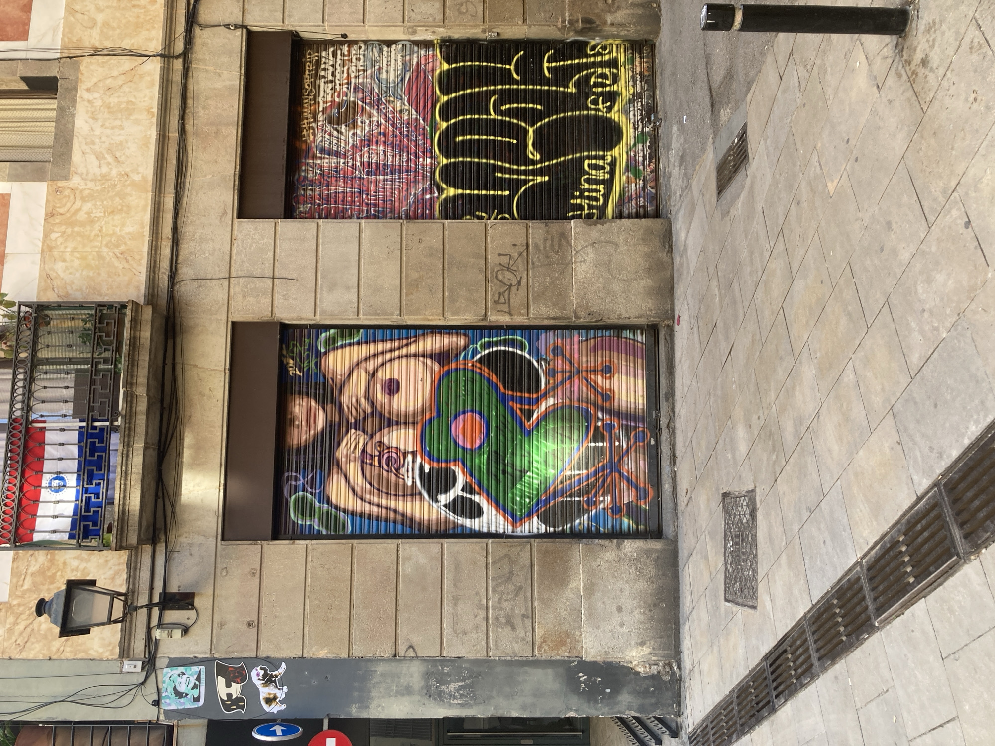 The image shows a street scene in Barcelona, Spain. Two window shutters are covered in graffiti. The top shutter has a colorful, chaotic design on the left side, with a black and yellow design on the right. The bottom shutter depicts cartoonish figures with a large green heart. The building facade is made of beige stone with visible seams. Electrical wiring runs down the side of the building. To the left, a window box containing a flag is visible. The sidewalk is paved with rectangular stones and has a long drain along its edge. A black post stands near the top right corner of the image. Stickers and a street sign are placed on the building's wall below the lower shutter.