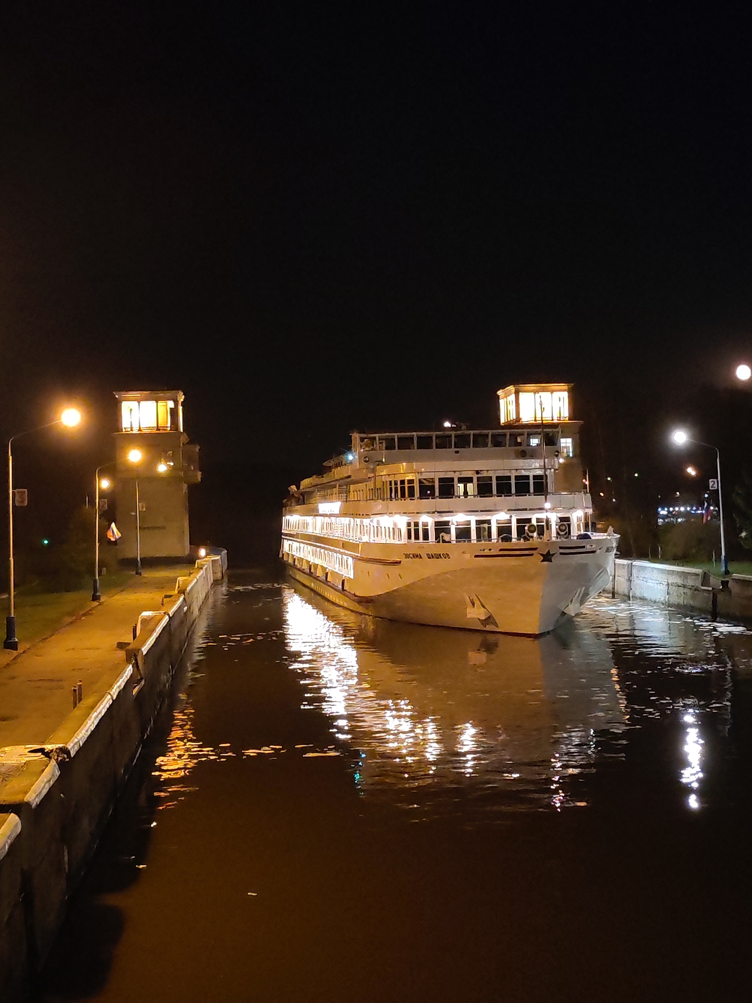 The image shows a river cruise ship, the "Josif Broz Tito", navigating a canal lock at night in Yakhroma, Russia. The ship, brightly illuminated with multiple rows of windows and a large star on its bow, is centered in the frame as it moves through the water. The reflections of the lights from the ship and surrounding structures are visible on the water's surface. The canal is flanked by concrete walls. On the left side of the canal, there is a building and a few street lamps. On the right, another structure and some greenery. The sky is dark, with a full moon visible in the upper right.