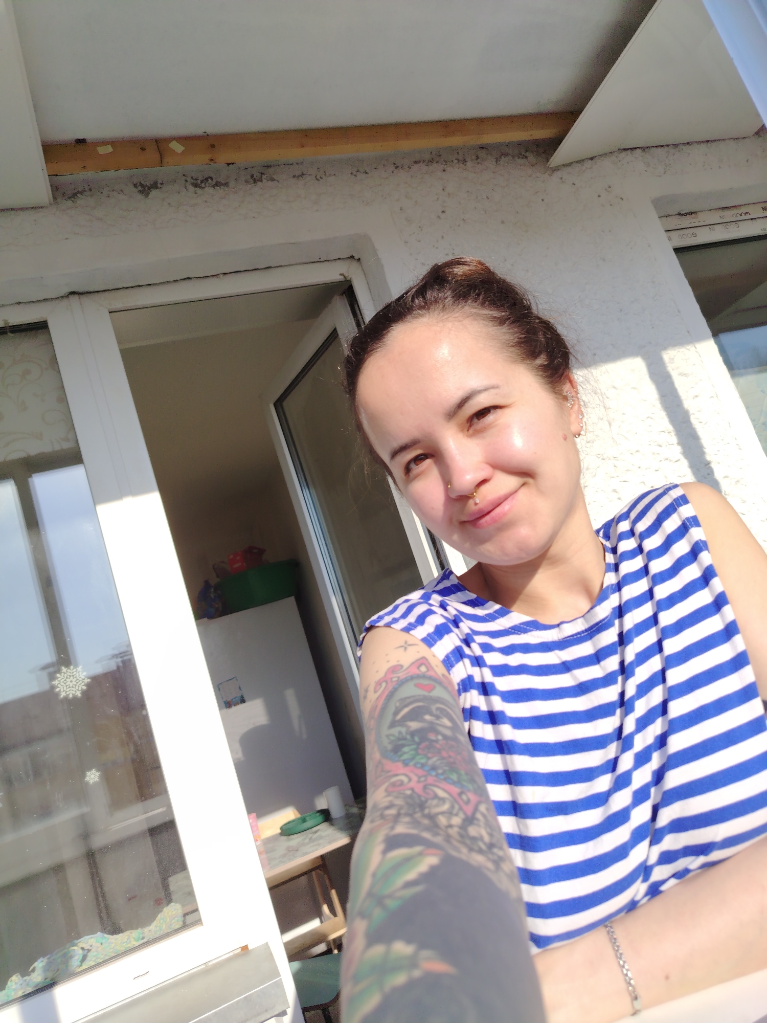 A woman is taking a selfie on a balcony. The woman has brown hair pulled back, a nose piercing, and is wearing a blue and white striped tank top. Her left arm is prominent in the foreground, displaying a colorful tattoo. A window with a snowflake sticker is to her left, leading to an interior space. Visible through the window is a refrigerator, a green bin filled with items, and a small table with a stool. The balcony has a white textured wall and ceiling. The location is likely Noviy, Russia, based on the user request, but this cannot be confirmed solely from the visual information. The lighting suggests it is daytime, with sunlight casting shadows.