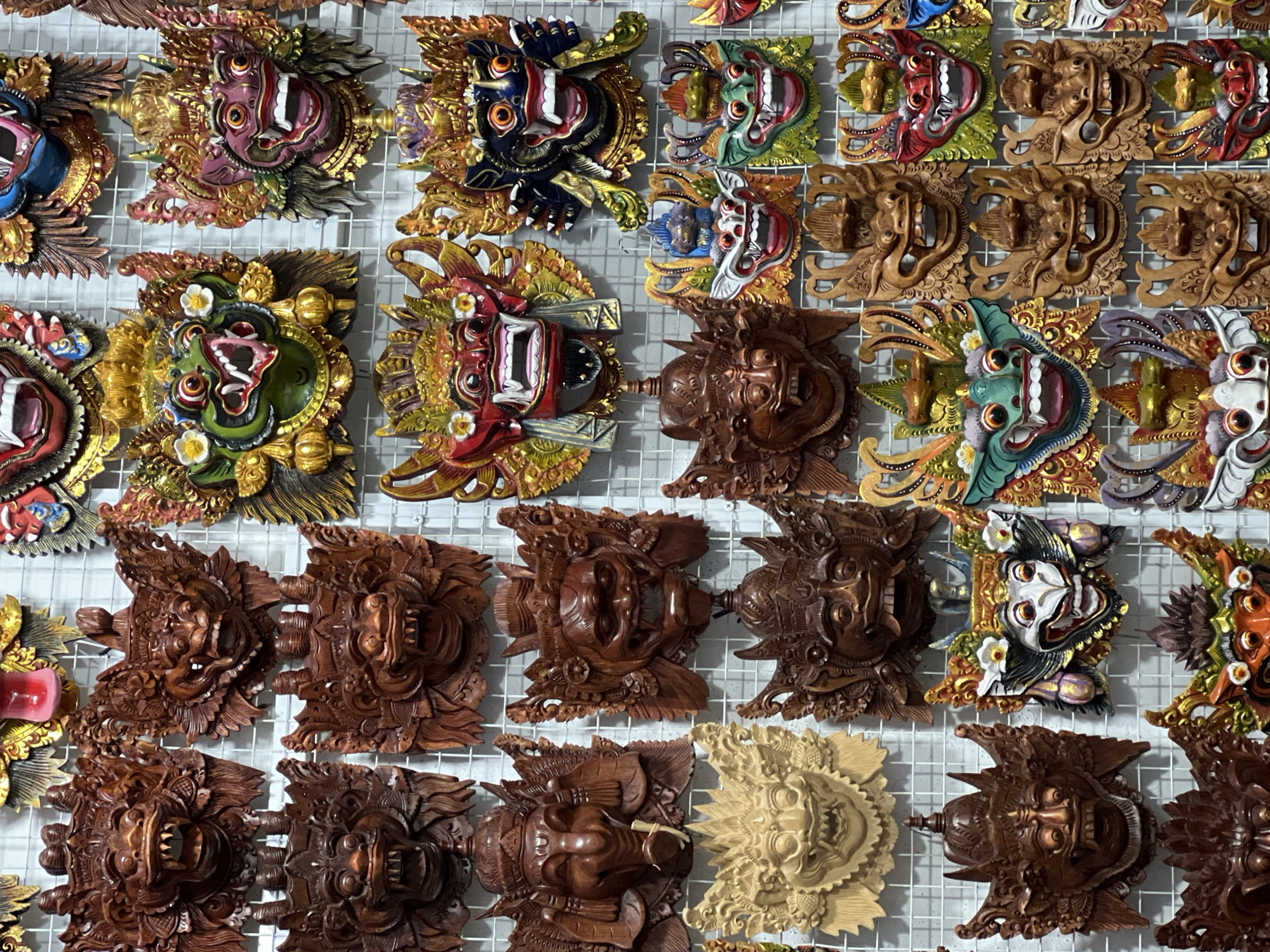 A colorful display of carved masks is arranged on a white wire grid, likely for sale. The masks vary in color and design, some are painted in vibrant shades with gold accents, while others are carved from wood in different tones of brown. All of the masks appear to depict mythical figures or deities, and they are the focus of the shot. The grid background indicates a possible market or shop setting. This image was taken in Ubud District, Indonesia.