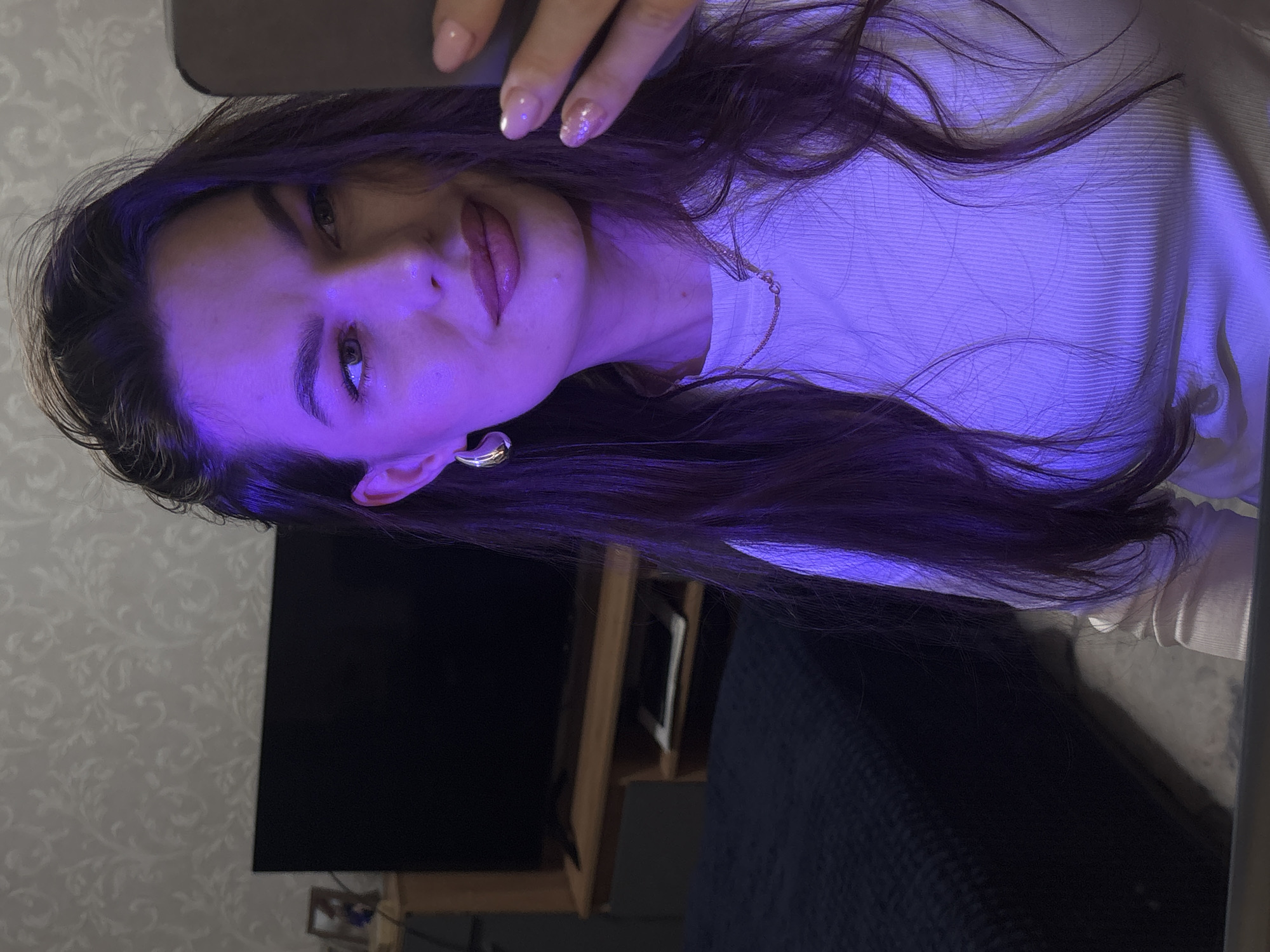 A young woman is depicted in a selfie, taken indoors, likely in a bedroom or living room in Chaikovsky, Russia. Her dark, long hair cascades around her shoulders and chest, partially covering a white ribbed long-sleeve top. A prominent purple/violet light source illuminates her face, hair, and upper body, casting a distinct glow and creating a moody ambiance. She looks directly at the camera with a neutral expression, her lips slightly parted.
Her right hand, featuring light-colored, manicured nails, is visible near her head, holding what appears to be the smartphone used to capture the image. She wears a gold chain necklace and a gold-toned hoop earring on her visible left ear.
In the background, a large, dark flat-screen television is visible to the left, positioned on a low wooden shelf or stand. The wall behind her is adorned with a light grey wallpaper featuring a subtle, ornate floral or damask pattern. A dark, textured fabric, possibly a bedspread or blanket, occupies the lower right portion of the frame. The artificial lighting suggests the photograph was taken during evening or night hours. No visible text is present in the image.