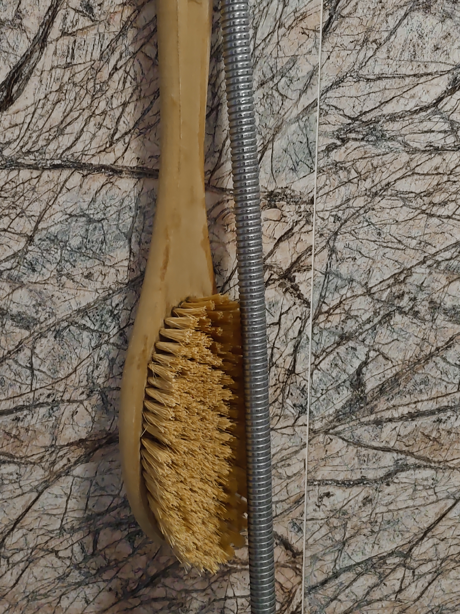 The image shows a close-up view of a tan-colored bath brush with natural bristles hanging next to a corrugated metal shower hose. The brush is positioned vertically, with its bristles facing towards the right side of the frame. The shower hose is also vertical, positioned to the right of the brush. Both items are set against a background of marble-patterned tiles, featuring a light beige base with intricate dark gray veins that resemble tree branches. The lighting is even, and there are no visible people or other objects in the frame. The image appears to be taken in a bathroom environment.