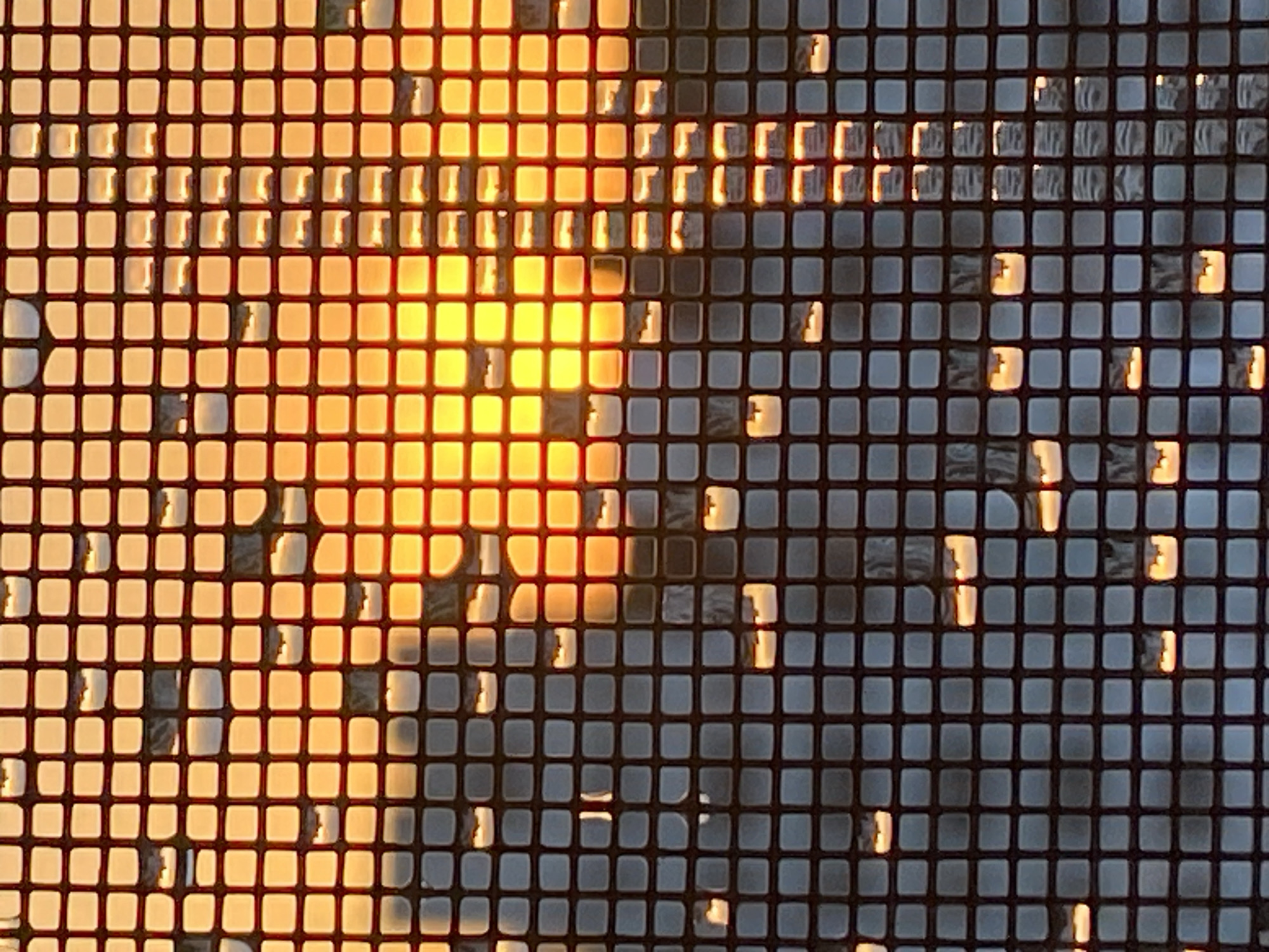 A close-up, abstract photograph captures the effect of sunlight filtering through a screen door. The screen is composed of a grid of small, square openings, each of which is illuminated or obscured by the light. The dominant colors are a vibrant orange and yellow from the sun, interspersed with darker, muted grays and blues that represent the shadows and the screen material itself. The light creates a shimmering, almost pixelated effect, with bright squares of light and darker areas where the screen is more opaque. Some of the illuminated squares have a soft, diffused quality, while others are sharper and more defined, suggesting variations in the screen's texture or the angle of the light. The overall impression is one of abstract beauty, focusing on the interplay of light and shadow on a textured surface. The location is not discernable from the image.
