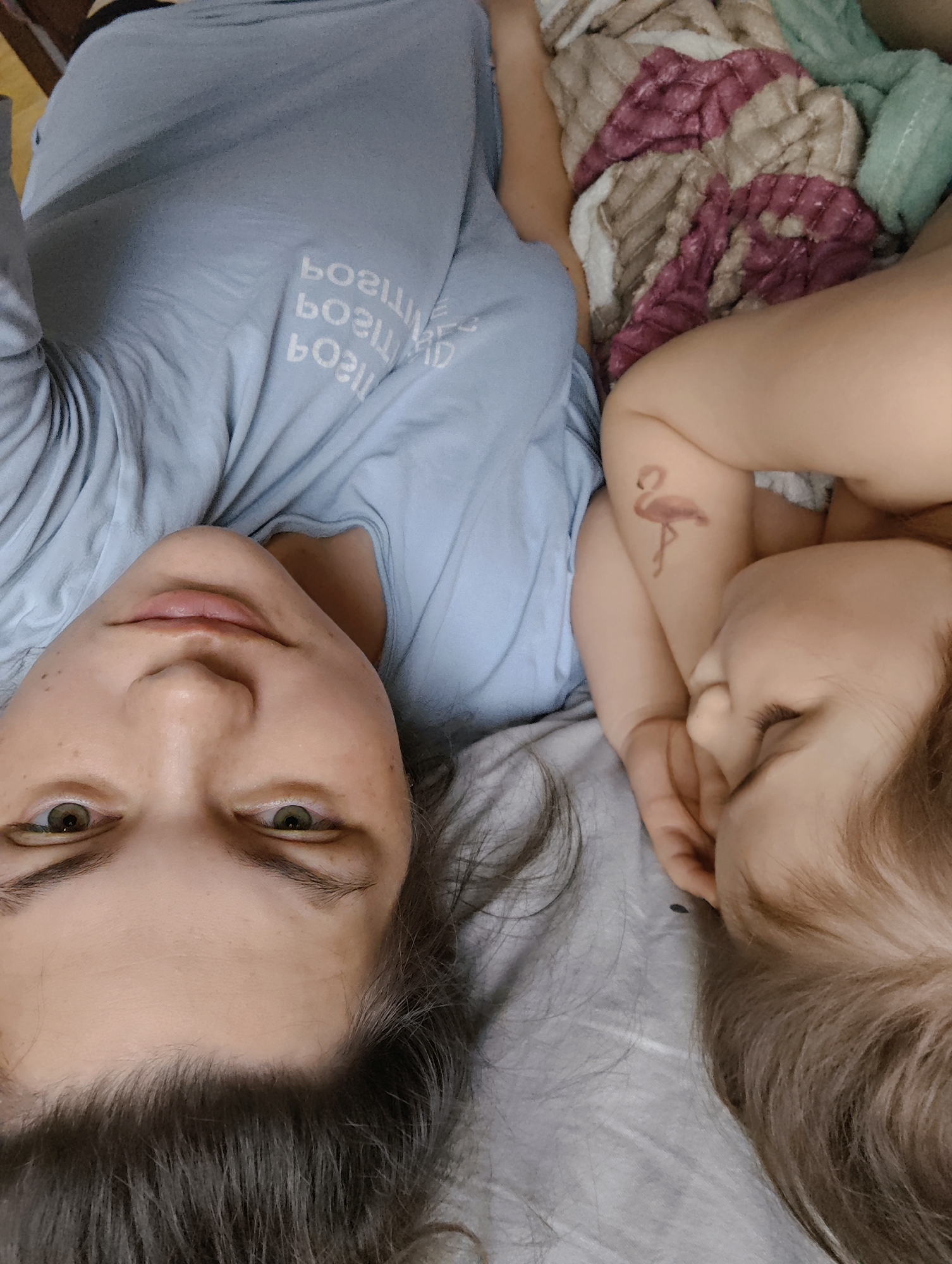 This vertical photograph captures an intimate indoor scene featuring an adult and a young child lying down, viewed from an overhead perspective. The setting appears to be a bedroom, likely on a bed, with light grey bedding and a textured blanket with beige, dark pink, and green patterns.
On the left, an adult, likely female, is positioned on their back, looking directly at the camera with a neutral expression. They have dark hair, green or hazel eyes, and a light complexion with visible freckles. The adult is wearing a light blue long-sleeved top. White text, mirrored and partially obscured by fabric folds, is visible on the shirt, which reads "РОЗИТИВ" (Russian for "POSITIVE") repeated several times.
To the right, a young child is nestled beside the adult, lying on their side with eyes closed, suggesting they are sleeping or resting peacefully. The child has light brown or blonde hair. A notable detail is a temporary pink flamingo tattoo visible on their upper right arm.
The lighting is soft and natural, contributing to a serene and calm atmosphere. The image portrays a quiet moment of connection or rest between the two individuals. The context of the photo is Chaikovsky, Russia.