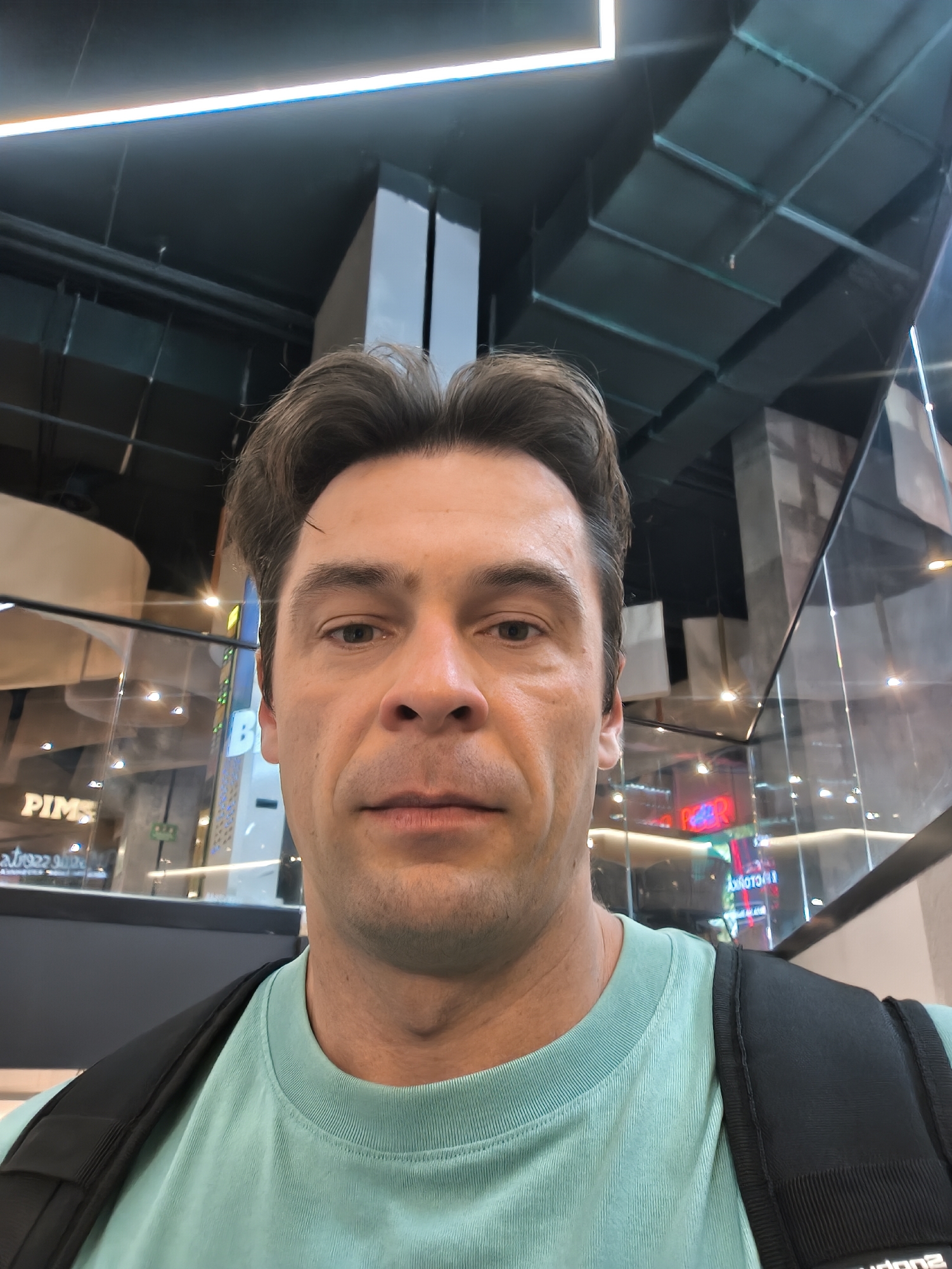 A medium-shot selfie of a middle-aged man with short brown hair and a light complexion wearing a light green t-shirt and a black backpack. The background shows an interior with retail spaces, glass walls, support columns, and overhead lighting. The retail sign "PIMS" is visible, as is the sign "FACTORY". The scene takes place within the VTB Arena in Moscow, Russia.