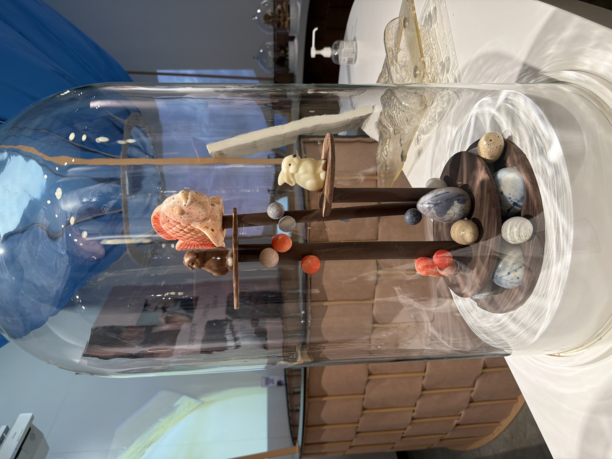 An elaborate culinary or artistic display is presented under a large, clear glass cloche, resting on a white surface. Inside, a multi-tiered structure of dark, vertical supports holds various sculpted pieces.
On a higher tier, a creamy white, plump bunny figure, possibly made of white chocolate, is perched. Below it, a reddish-pink, textured, shell-like or abstract piece is positioned. Further down, a smaller, dark brown, animal-shaped figure is visible. Scattered throughout the structure and on the base are numerous small, irregularly shaped items resembling eggs or pebbles, in speckled beige, blue-grey with white marbling, solid red-orange, and light beige.
The glass cloche reflects its surroundings. To the left, a vibrant blue fabric, possibly a curtain, is visible. On the right, the white table surface extends, holding a hand sanitizer dispenser with "Hydro alcoholic gel" partially visible, and delicate, translucent decorative pieces, including another smaller glass dome. Reflections of a light-brown, brick-like wall pattern and a blurred projection screen are also evident.
The scene suggests an exhibition or a sophisticated dessert presentation in a public setting in Illkirch-Graffenstaden, France. The items appear whimsical and meticulously crafted.