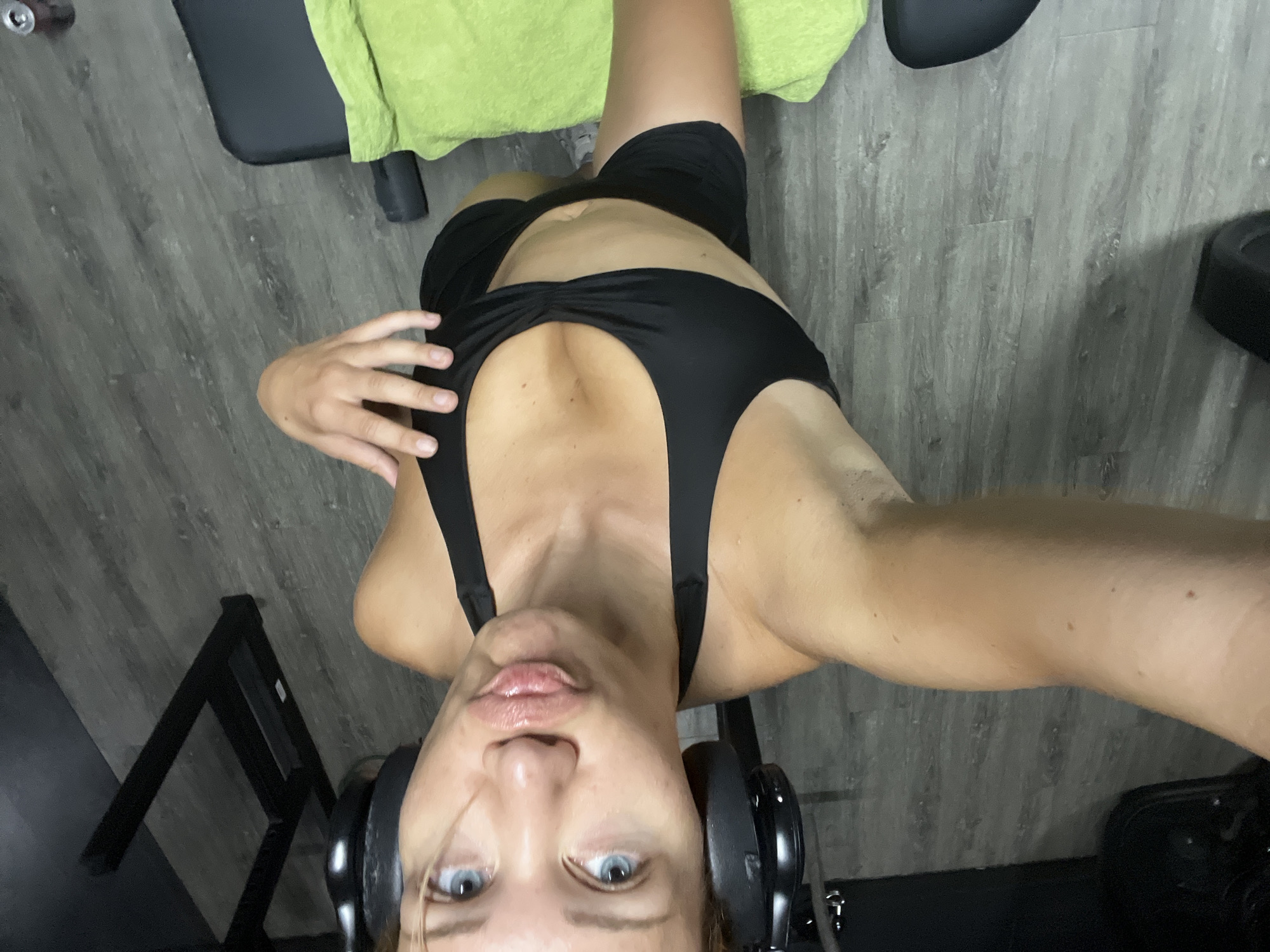 A woman wearing a black sports bra and black shorts, with black headphones on, takes a selfie in what appears to be a gym. She is looking at the camera with her lips pursed and has her left hand on her chest. A green towel is visible on a black bench or machine in the foreground. The floor is a grey wood-like material, and gym equipment is visible in the background. The image does not contain any elements that would place it in the city of Altötting, Germany.