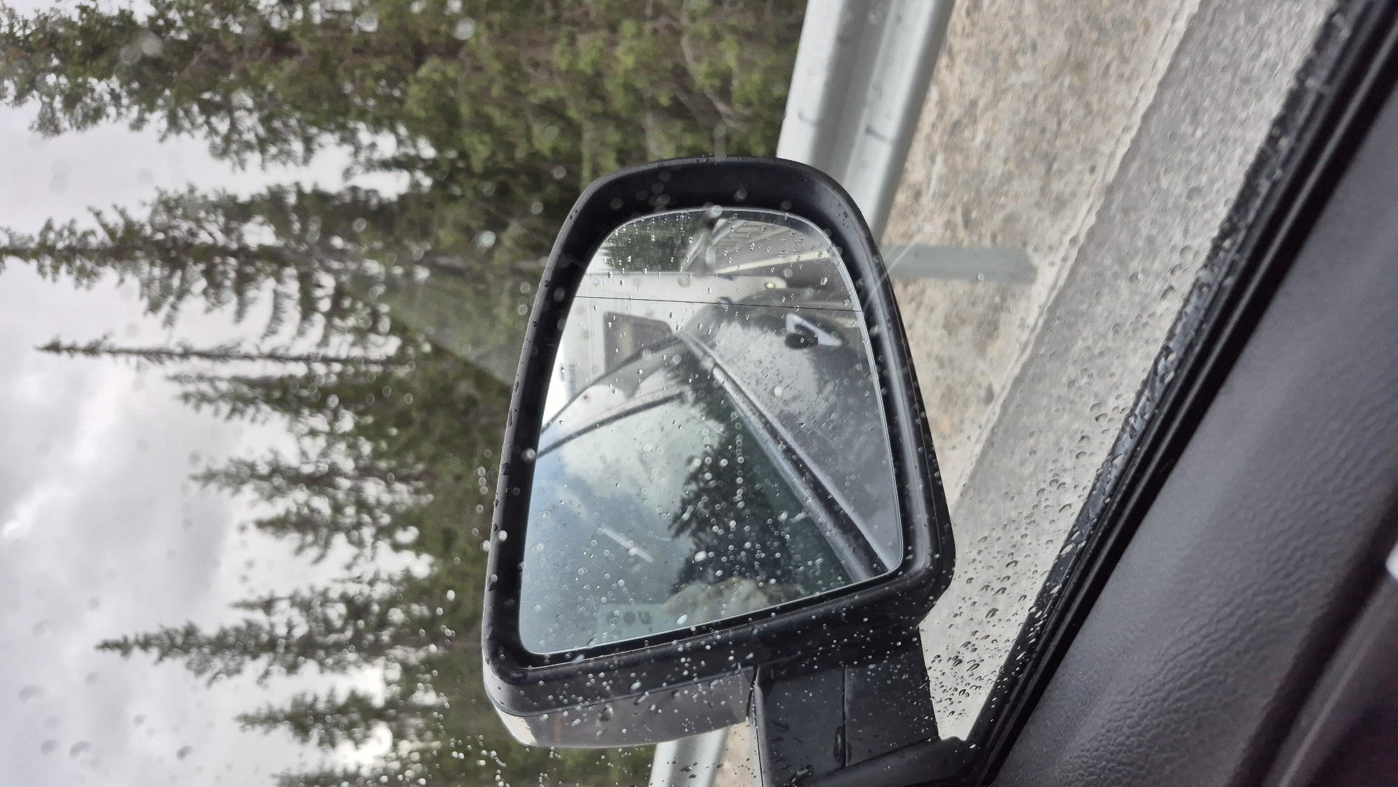 The image shows a close-up view of a car's side mirror during a rainy day, reflecting the surrounding environment. The mirror itself is speckled with raindrops, and the housing is dark gray. In the reflection, the side of another vehicle, possibly a truck or trailer, is visible, as well as a road and a guardrail. Behind the road, a dense forest of evergreen trees is present under a cloudy sky. The interior of the car, specifically the door panel, is visible in the foreground, also showing raindrops on the window. The location is identified as Nesland, Norway, suggesting a driving scene on a roadway in a forested area.