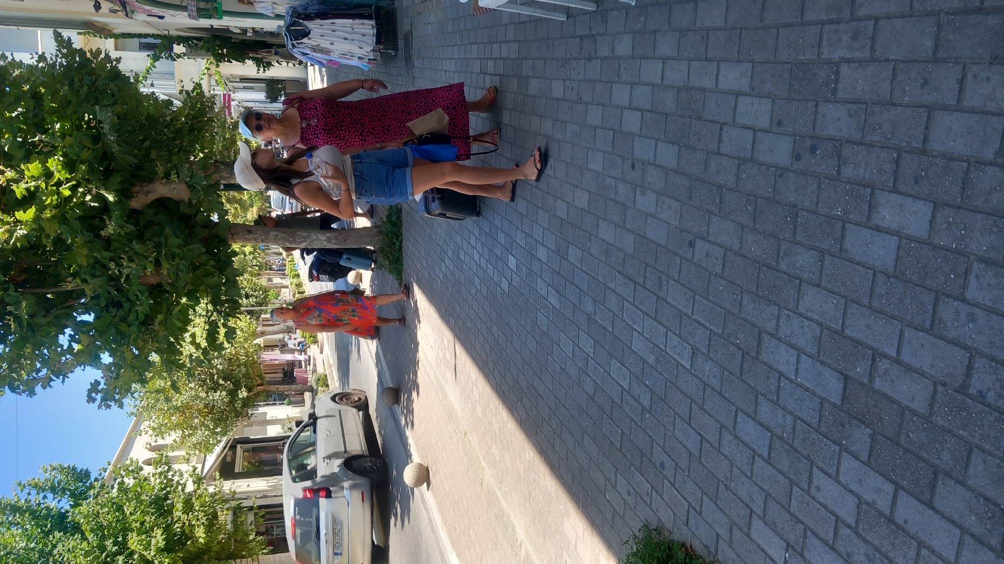 The image captures a street scene in Kos, Greece, under a bright, sunny sky. The foreground is dominated by a paved walkway made of grey rectangular bricks. A long shadow from a tree stretches across the walkway, dividing it into sunlit and shaded areas. To the left, a silver car is parked on the street, with a license plate beginning with "EX". Two round, concrete bollards are visible on the edge of the street.
In the middle ground, three women are walking on the sidewalk. The woman on the left wears a vibrant, orange and red patterned dress. She is walking away from the camera. In the center, a younger woman wearing a white baseball cap, a light-colored tank top, and denim shorts is walking towards the camera. She holds a water bottle and has a blue bag attached to her left side. Next to her, a woman with short grey hair and sunglasses wears a patterned pink dress. She is also walking towards the camera and carries a brown paper bag. Both women in the middle ground have luggage near them; the younger woman has a black rolling suitcase and a blue bag, while the other woman has a black suitcase.
Further down the street, more trees line the sidewalk, and buildings with architectural details are visible. A rack of clothes is displayed outside a shop on the right side of the image. The overall impression is a bustling urban environment on a warm day.