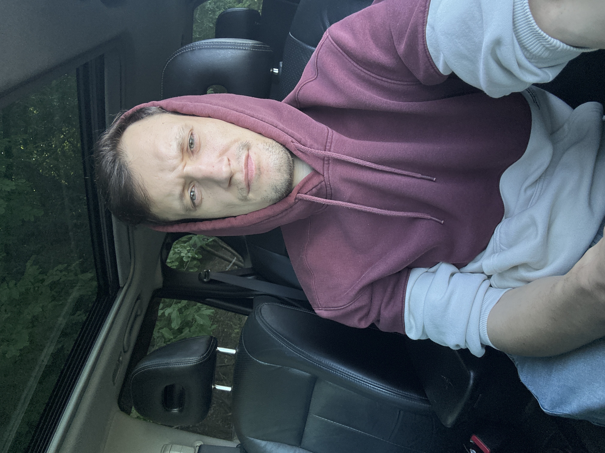 A man is sitting in the driver's seat of a car. He is wearing a maroon and white hooded sweatshirt. The car appears to be parked. The background shows trees and foliage, suggesting a natural or semi-rural environment. The image does not provide any specific visual cues that would place the location in the city of Makiv, Ukraine.