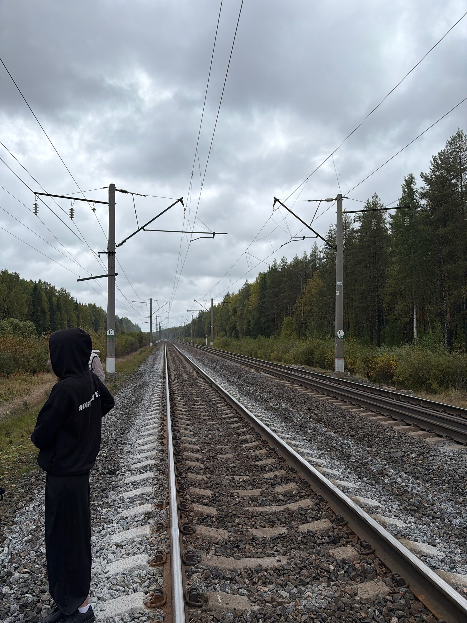In Kukhtoozerskiy, Russia, a person wearing a black hooded sweatshirt and black pants stands facing away from the camera, looking towards a set of train tracks that recede into the distance. The sweatshirt has white text on the back. The train tracks are flanked by a wooded area with a mix of deciduous and coniferous trees. Overhead, a complex network of electrical wires and poles associated with the railway system is visible against a cloudy sky. The ground around the tracks is covered in gravel and ballast.