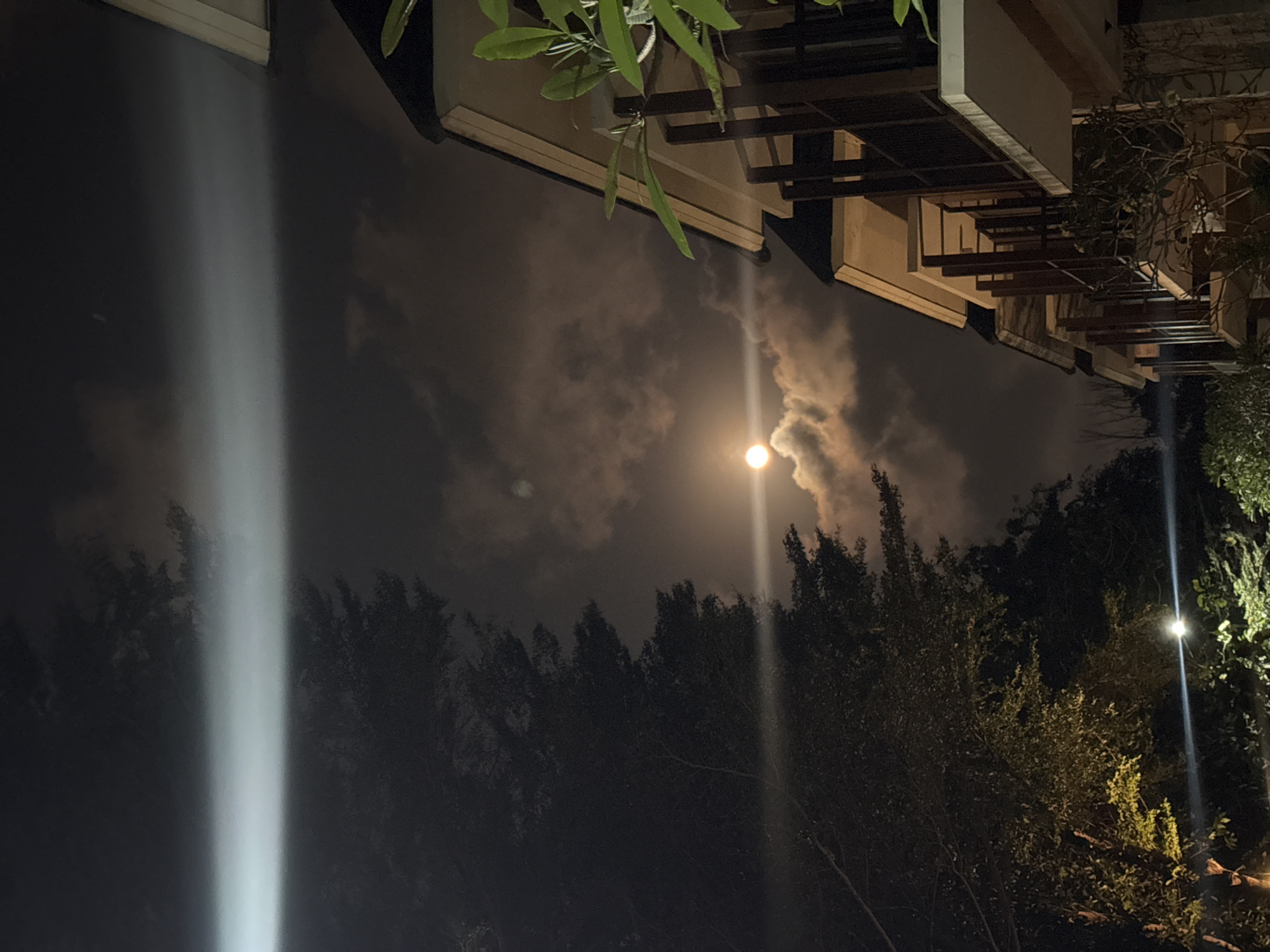 The image depicts a nighttime scene in Kuta Selatan, Indonesia, featuring a celestial event and architectural elements. A bright moon, partially obscured by clouds, is visible in the dusky sky, casting a warm glow. The clouds themselves are illuminated, creating a dramatic contrast with the darker sky. To the right, a modern building with multiple balconies and dark railings is partially visible, its facade illuminated by ambient light. A lush tree with dense foliage occupies the left side of the frame, its dark mass contrasting with the lighter sky and the building. A bright horizontal light beam cuts across the upper portion of the image, possibly from a distant light source or a lens flare. Another bright light source, possibly a streetlamp, is visible lower down in the frame, illuminating some of the foliage. The overall atmosphere is serene and atmospheric, capturing a tranquil night in a tropical setting.
