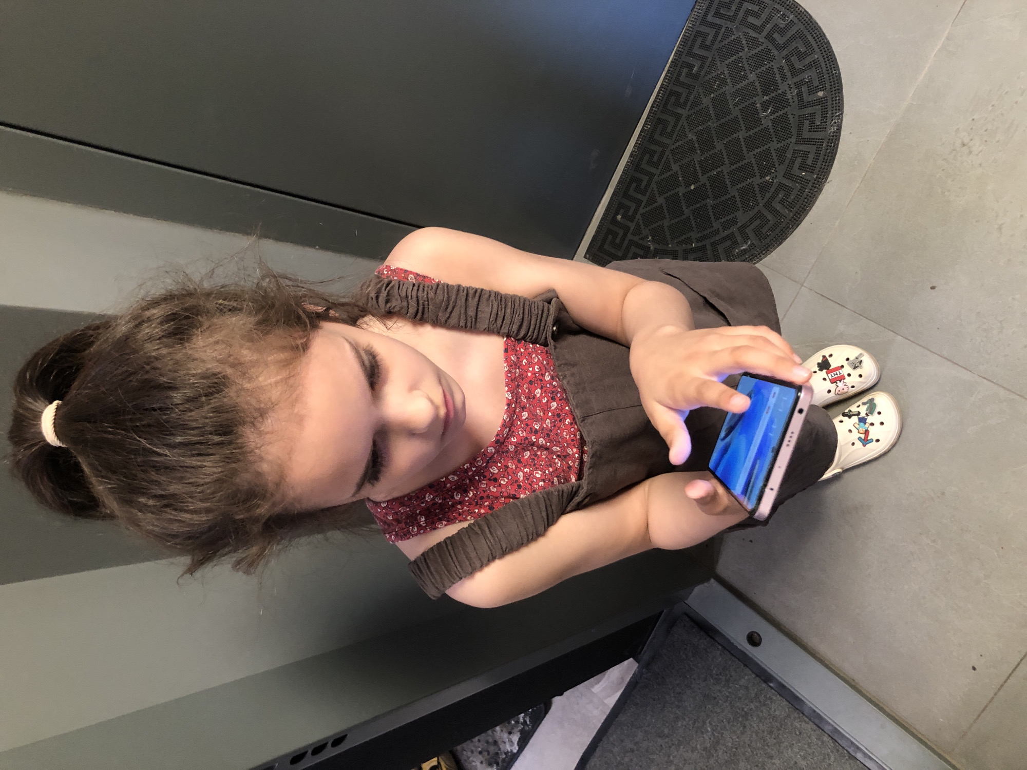 A young female, estimated to be between 8 and 12 years old, stands indoors in Saint Petersburg, Russia, concentrating on a smartphone. Her dark brown hair is pulled into a high, somewhat messy ponytail secured with a light-colored elastic band. She wears a dark brown pinafore dress with ruched shoulder straps over a red top featuring a dense pattern of small, light-colored floral or abstract shapes. She holds a light-colored smartphone horizontally with both hands, actively interacting with the illuminated blue-toned screen using her right index finger. Her feet are clad in white Crocs-style clogs; the left clog displays multiple colorful charms, including what appears to be a green pipe, a red mushroom, and a red "TNT" block, while the right clog features at least one red, pig-like charm. She stands on a light grey tiled floor. The background consists of two distinct vertical wall panels: a lighter, greenish-grey panel to the left and a darker grey panel to the right, separated by a thin vertical line. A dark grey, ridged mat is partially visible on the far left, and a semi-circular dark rubber mat with a repeating geometric pattern is situated on the floor to the far right, bordering the darker wall panel.