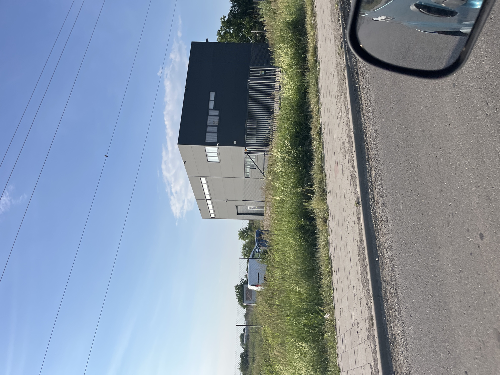 The image depicts a modern, dark gray, rectangular building with multiple windows, situated in a grassy, semi-rural area. The building appears to be an industrial or commercial facility. Several vehicles, including a white van and a dark-colored SUV, are parked in front of the building behind a metal fence. Tall grass and some trees surround the property. Power lines are visible stretching across the bright blue sky, which has a few wispy clouds. A sidewalk runs along the edge of a paved road in the foreground, and the reflection of a light blue vehicle is visible in a car's side mirror, suggesting the photo was taken from a moving vehicle. The scene is likely located in Mynai, Ukraine, given the context.