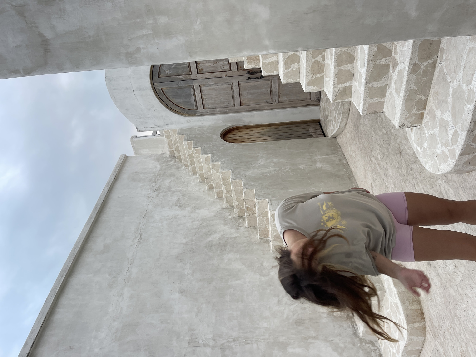 A person with long, dark hair is captured mid-motion, shaking their head in an outdoor courtyard in North Kuta, Indonesia. The individual is wearing a light grey t-shirt with a yellow graphic and pink shorts. They are standing on a textured stone floor, with a staircase constructed of similar stone to their right, leading upwards. The surrounding architecture is characterized by smooth, light grey concrete walls with a textured finish. Behind the staircase, a large, arched wooden door is visible, set into a curved wall. To the left of the main staircase, a smaller, arched wooden door is also present. The sky above is overcast with clouds.