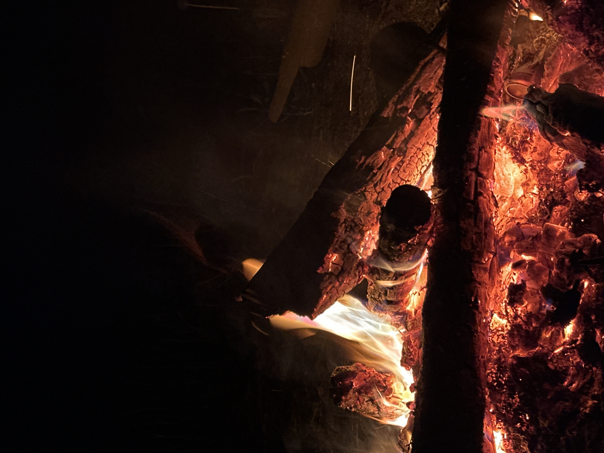 A nighttime image shows a close-up of a wood-burning fire. The fire consists of stacked logs and glowing embers. Bright orange and white flames emerge from the center of the fire. The surrounding area is dark, with some indistinct shapes and objects visible. No people are visible in the image. It is not possible to determine whether the image was taken in Юзуфоўскі сельскі Савет, Belarus.